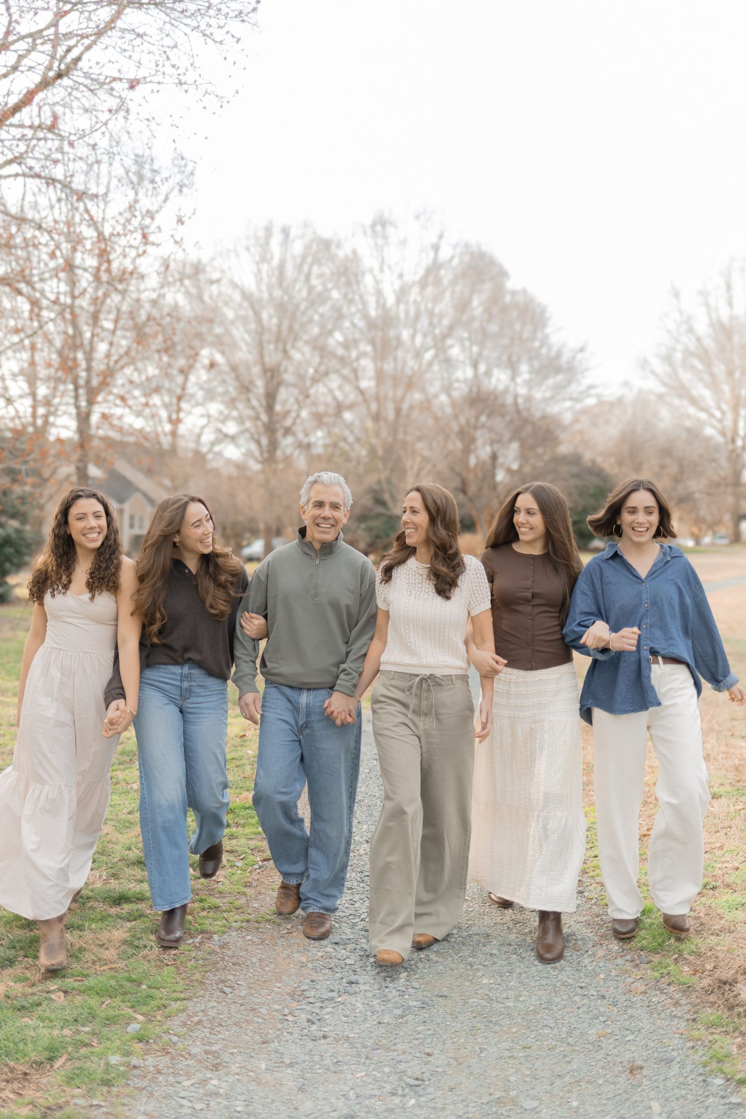 A joyful family of six walks together on a gravel path outdoors, dressed in neutral tones during a fall family portrait session.