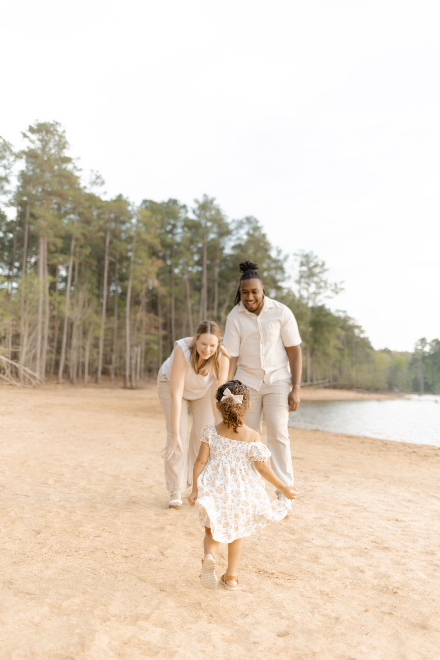 Family photo on sandy beach with tall pines, parents in neutral tones welcoming young daughter in white dress.