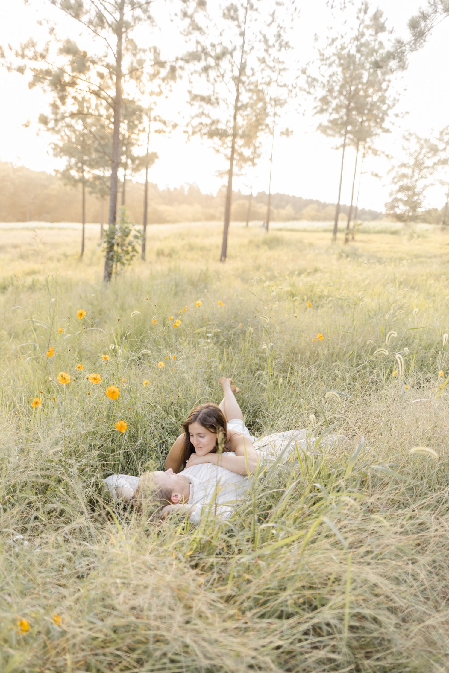 A young woman in a white dress reclines in a sunlit meadow with wildflowers and tall pine trees in golden hour light.