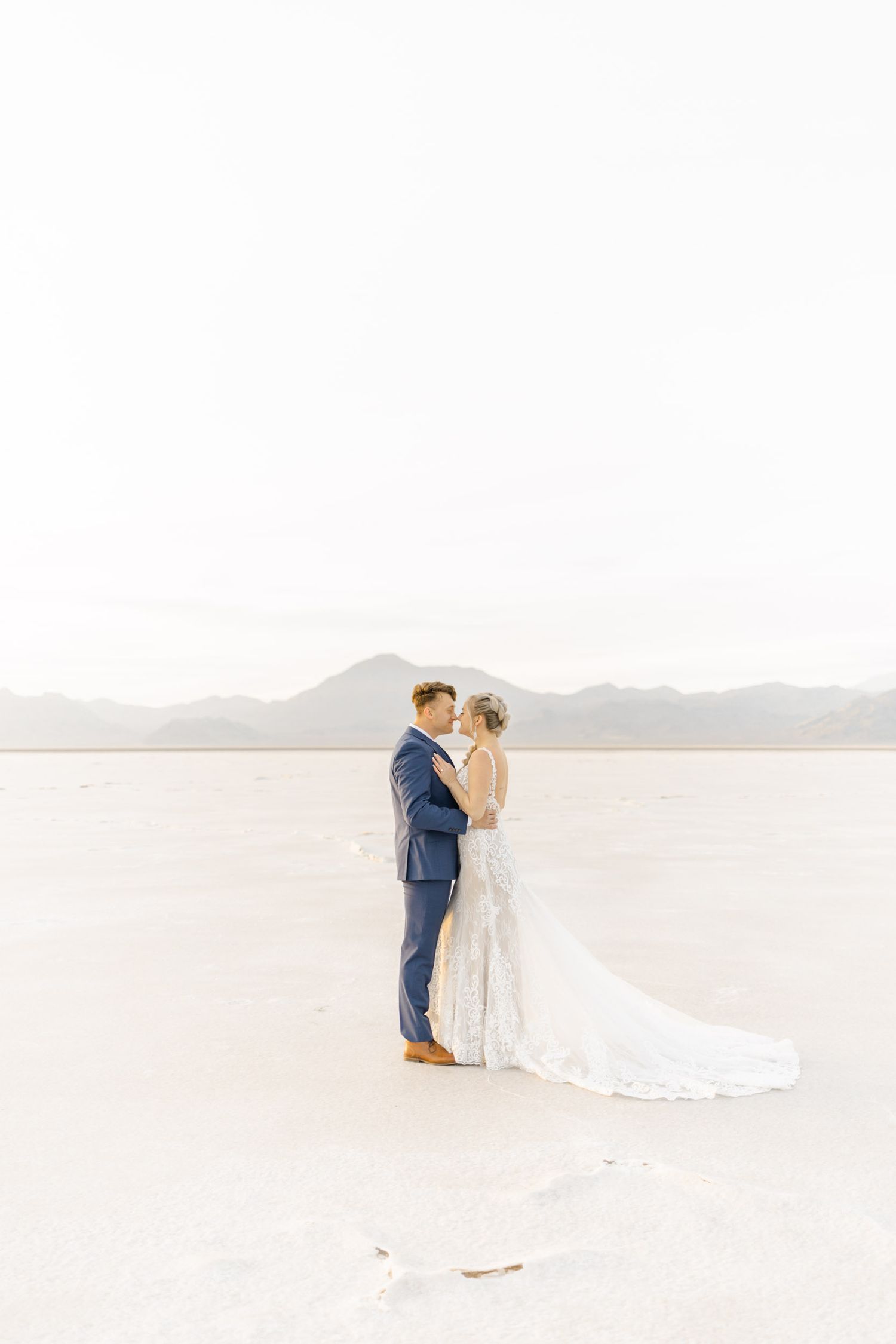 A couple shares a romantic moment in white wedding attire against a serene desert landscape with mountains in the distance.