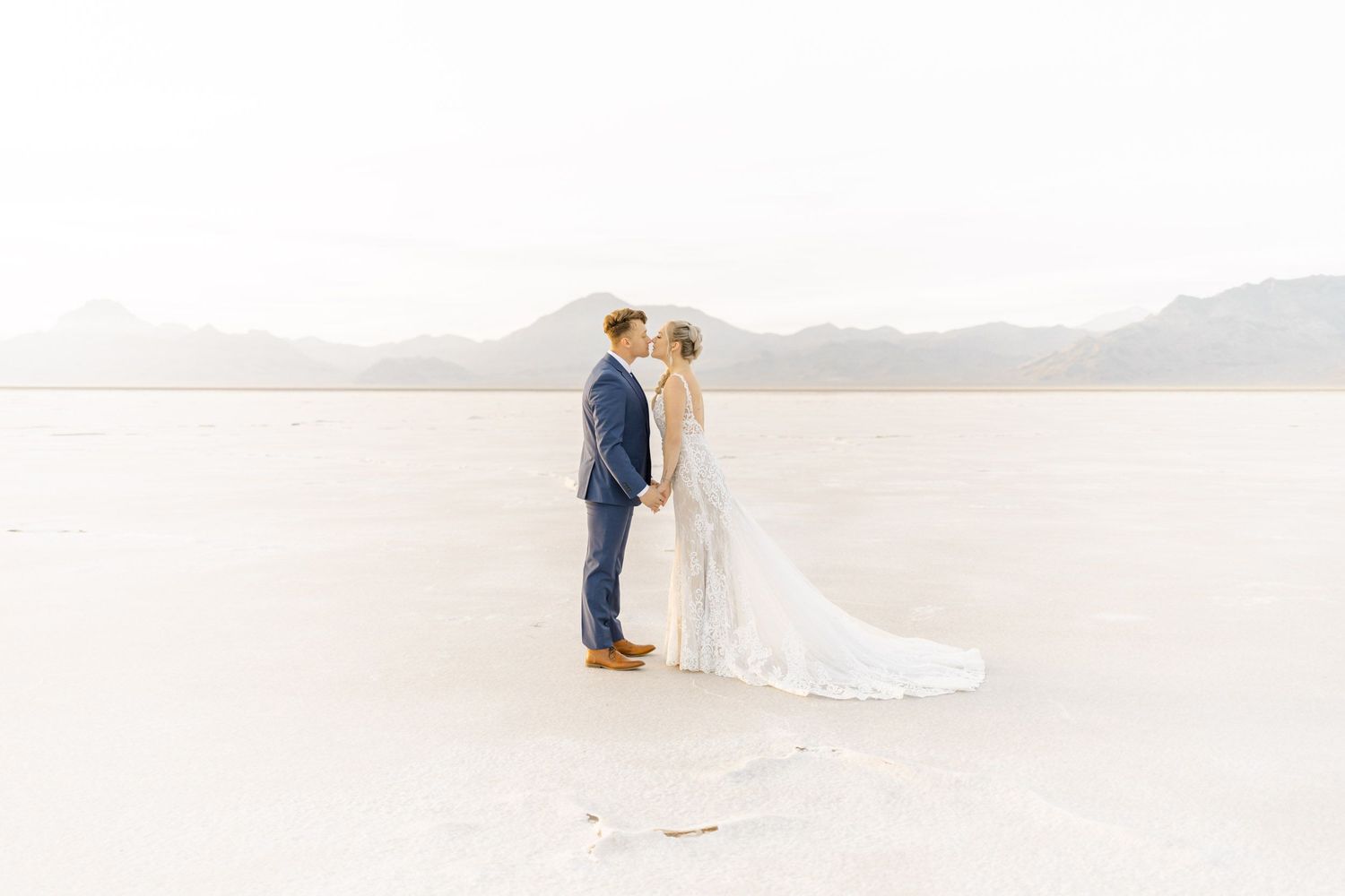 A couple stands together on a white salt flat landscape with mountains in the distance.