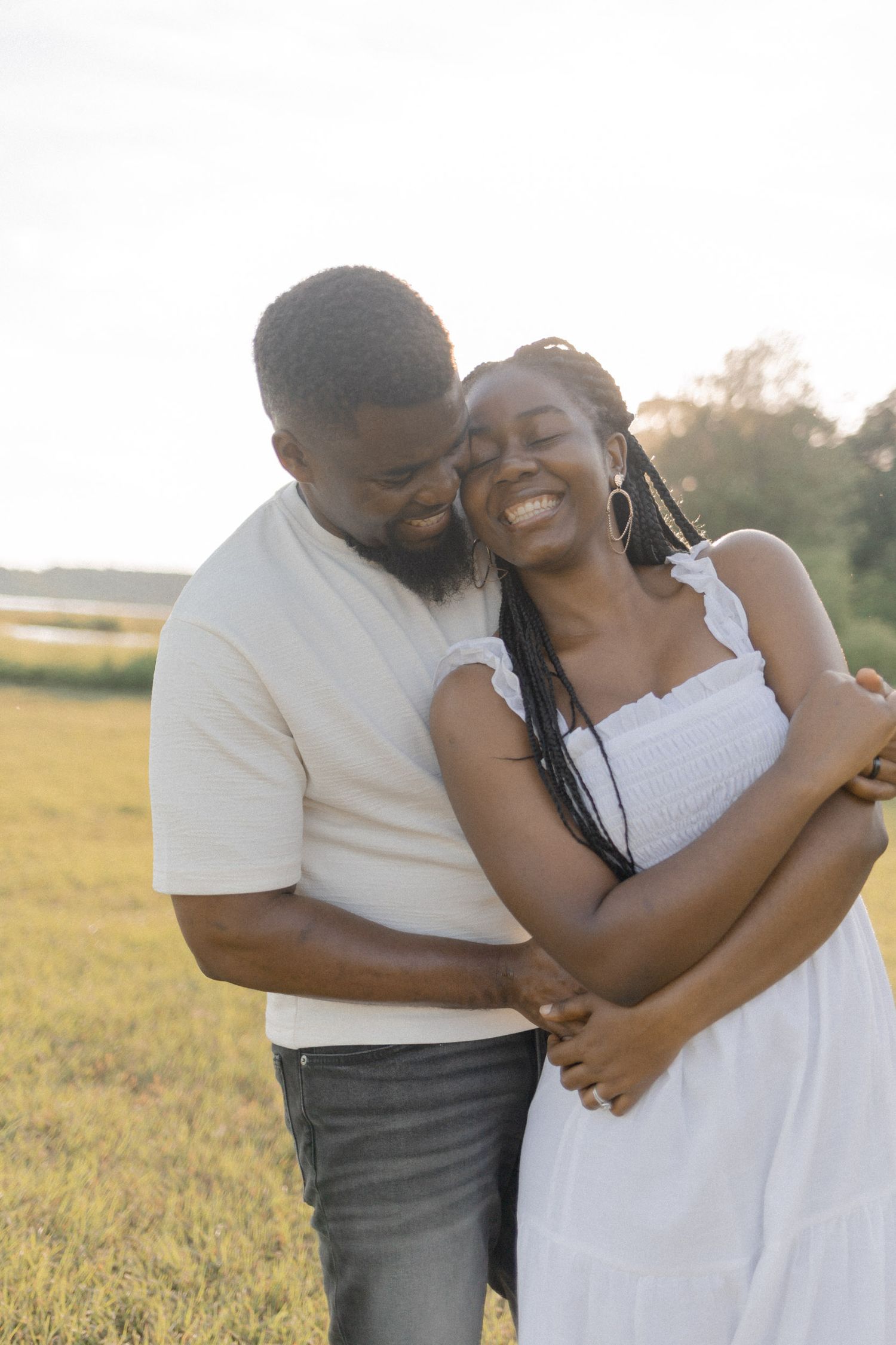 A happy Black couple embraces and laughs together in a sunny outdoor field, woman in white dress, man in cream shirt.