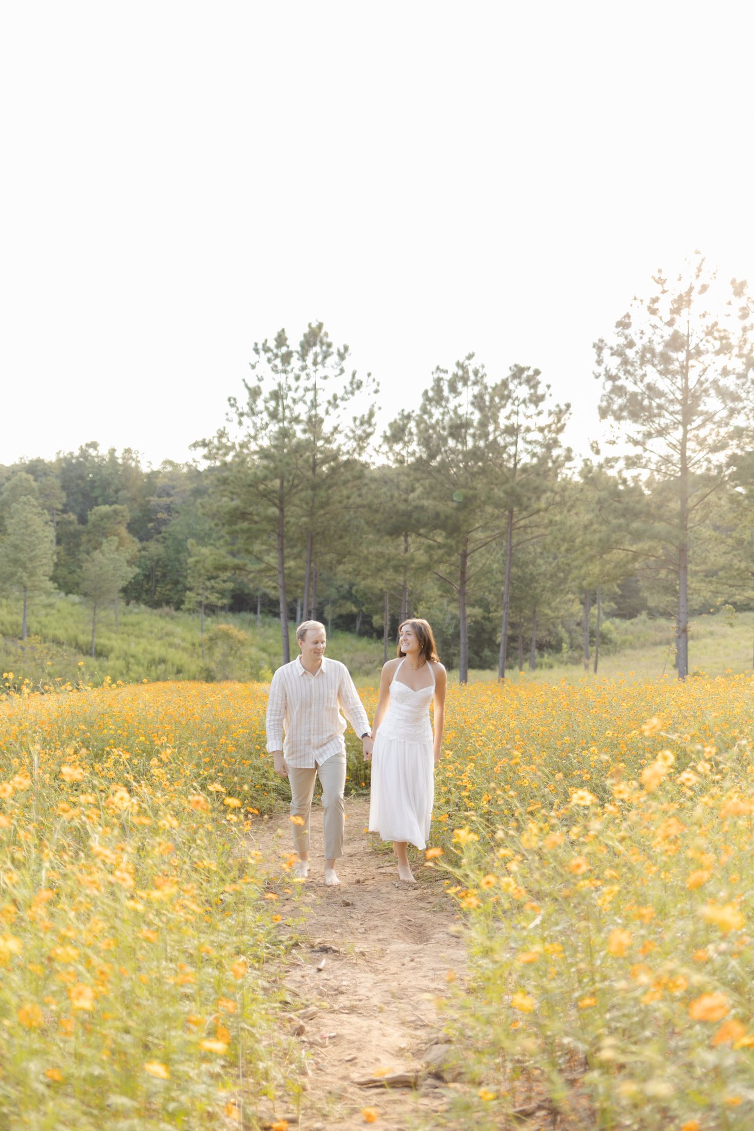 Couple strolling down a dirt path through a meadow of yellow wildflowers at sunset during lifestyle portrait photoshoot.