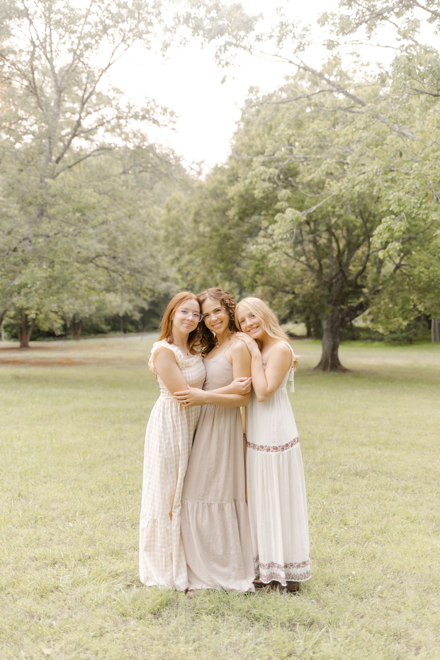 Three women in flowing neutral dresses share a group hug outdoors in a pastoral setting with Durham, North Carolina Photographer.