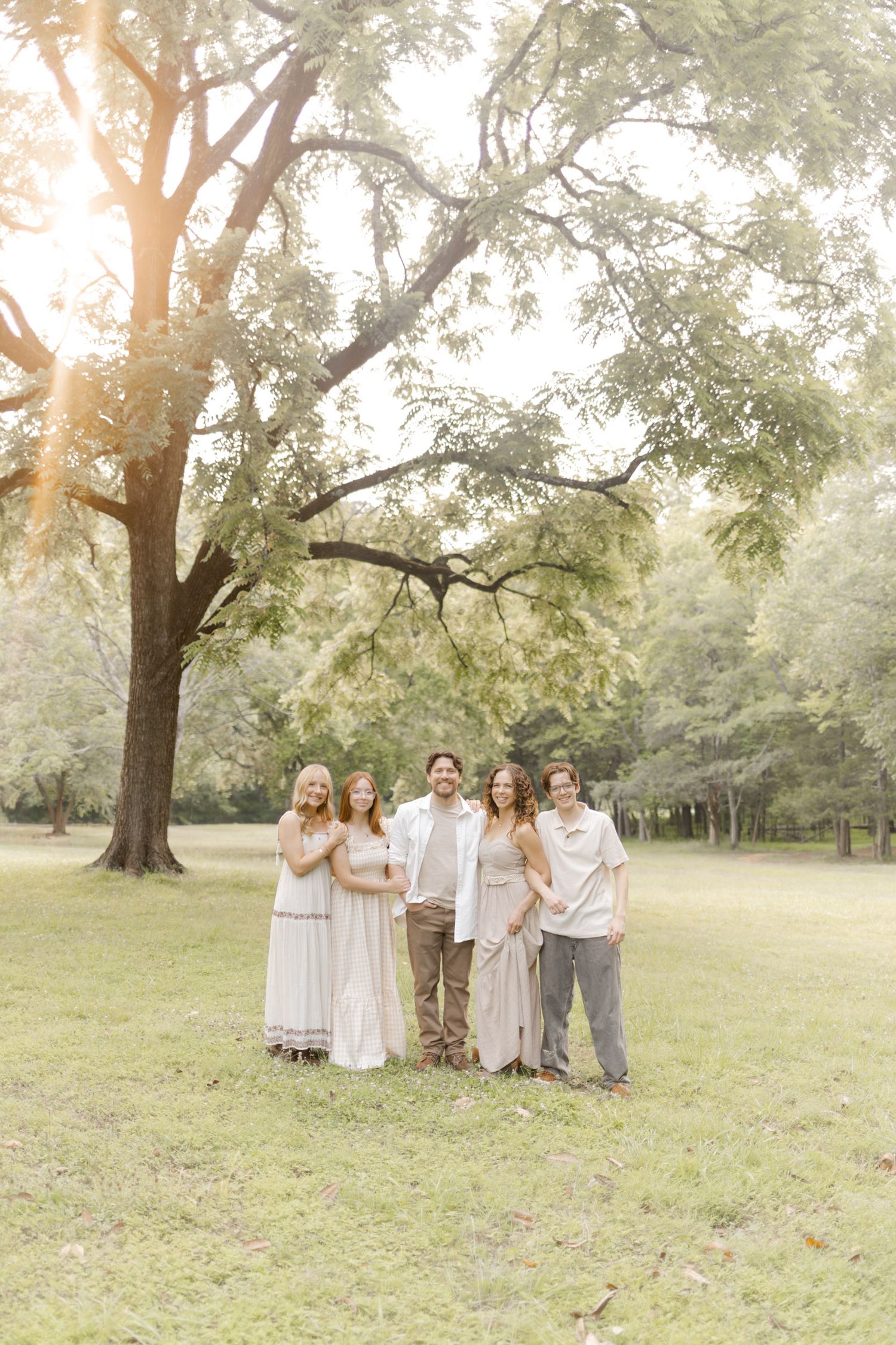 Family group poses for photos in a sunlit park wearing coordinated neutral-colored clothing while doing a photoshoot.