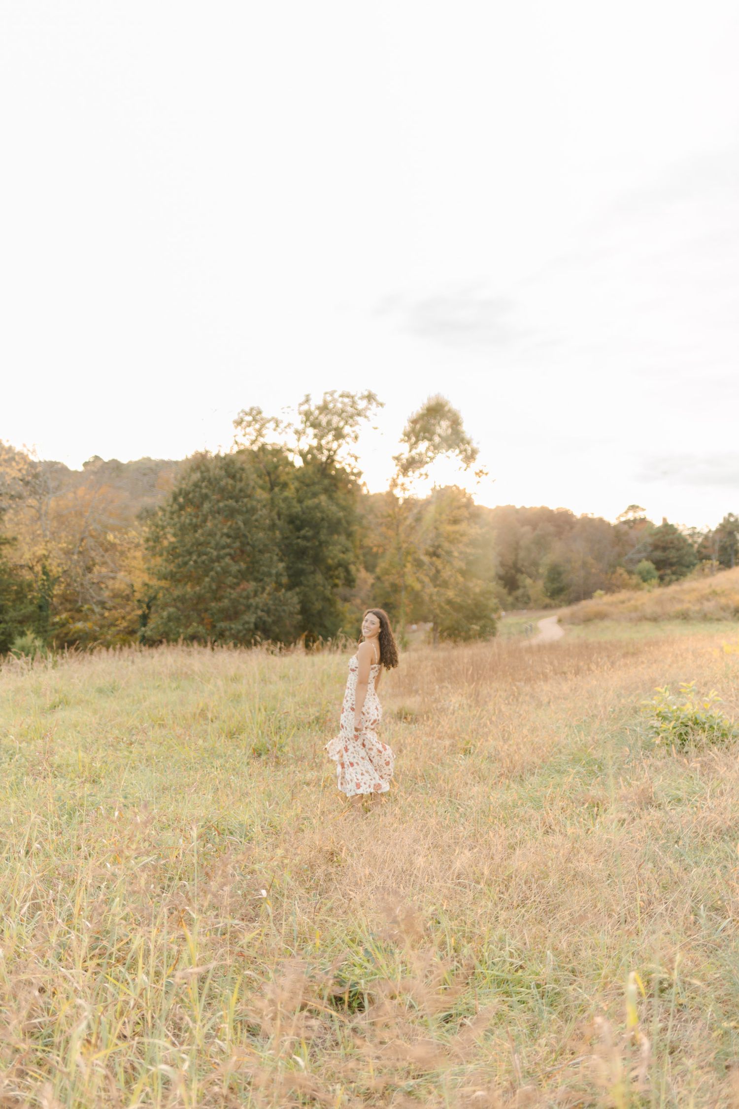 Young woman in floral dress standing in sunlit meadow surrounded by autumn trees and golden grass.
