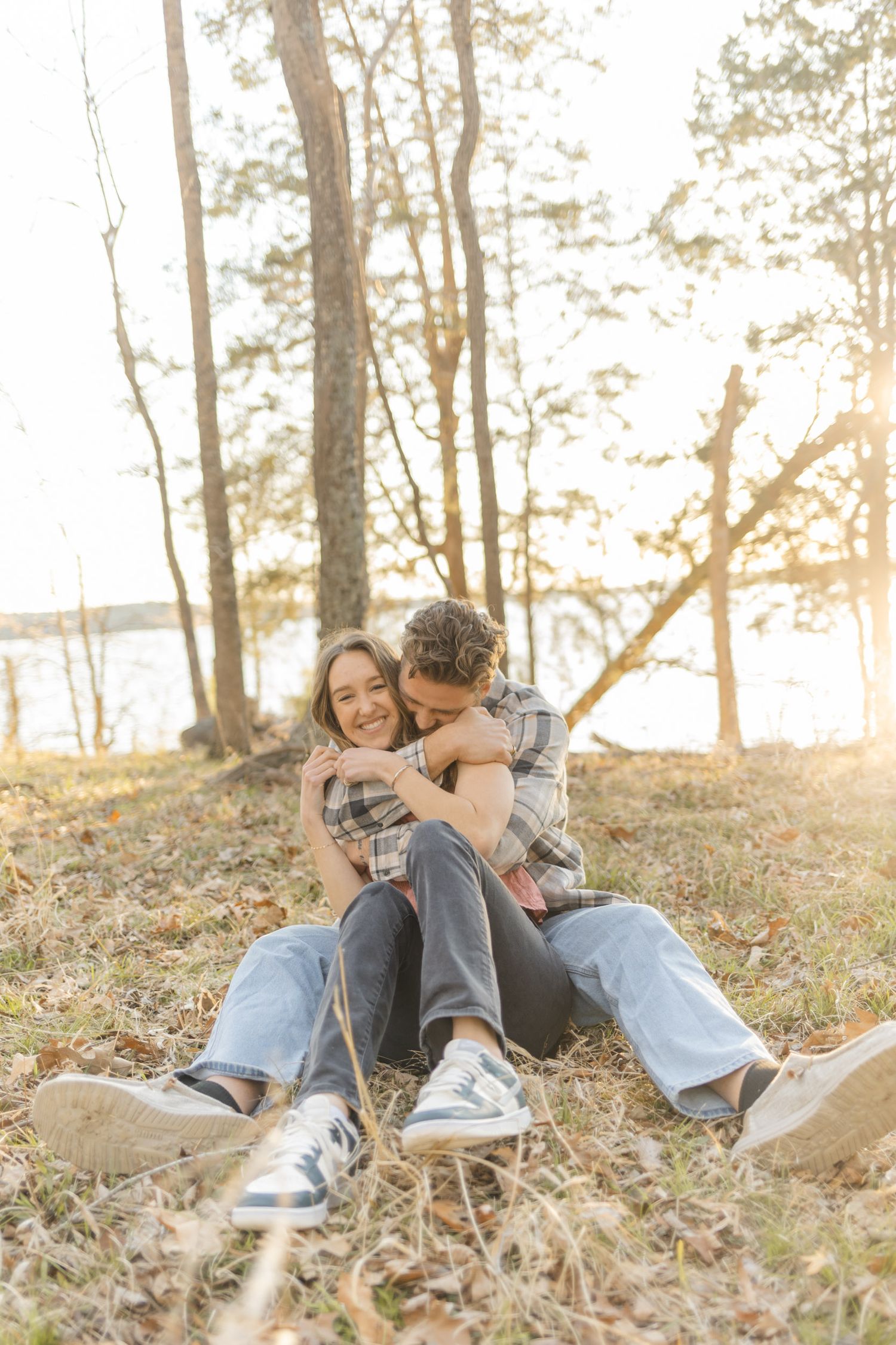 Couple sitting together on autumn leaves in a sunlit forest, sharing a tender embrace near a serene lakeside.