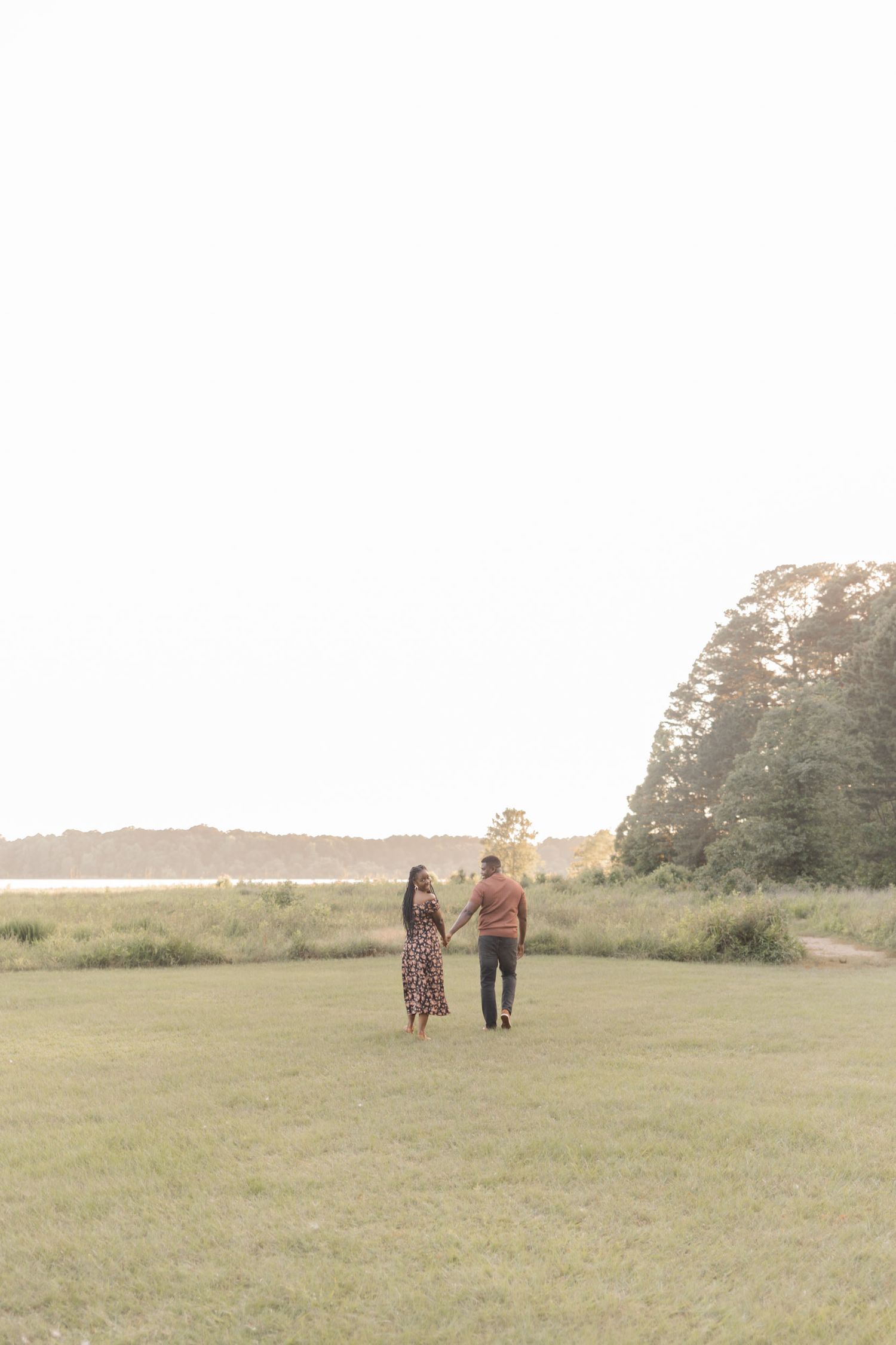 Couple holding hands walking through a green field toward a serene waterfront surrounded by trees at golden hour.