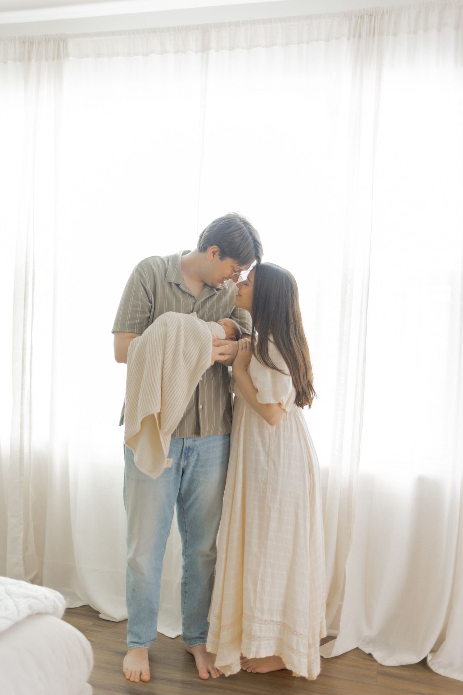 Couple sharing a tender kiss while holding their newborn baby by a bright window.
