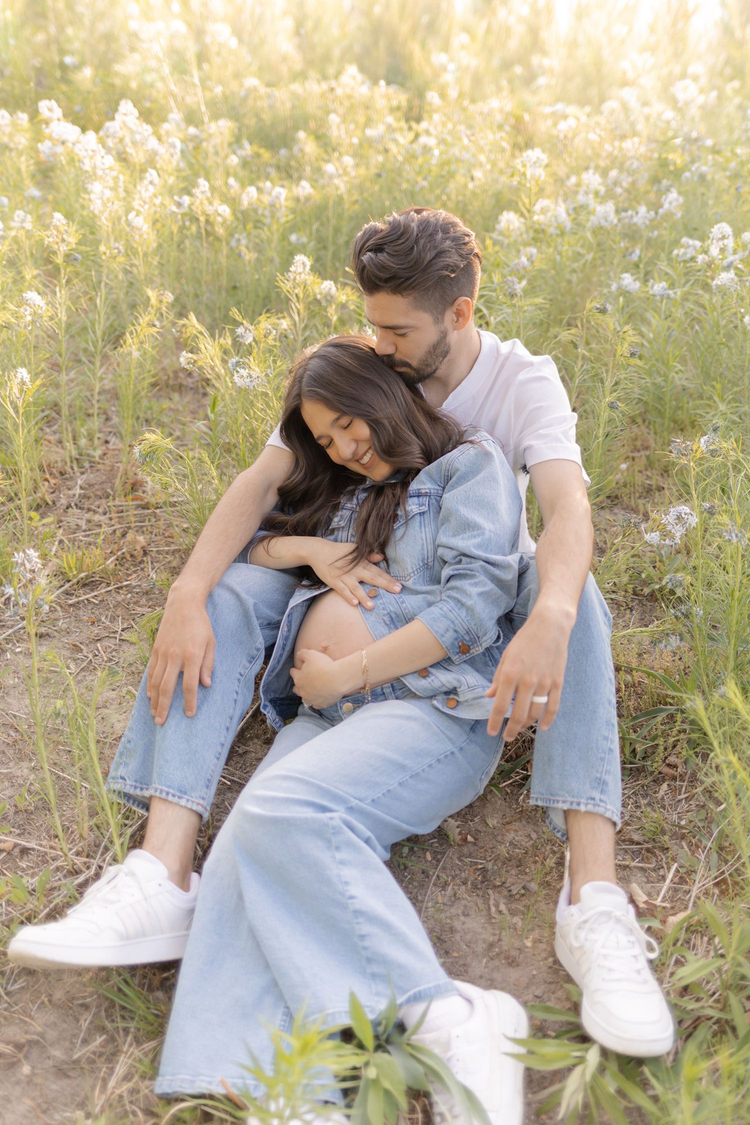 Pregnant couple sitting in flower field at sunset, man kissing womans head while cradling baby bump in denim outfits.