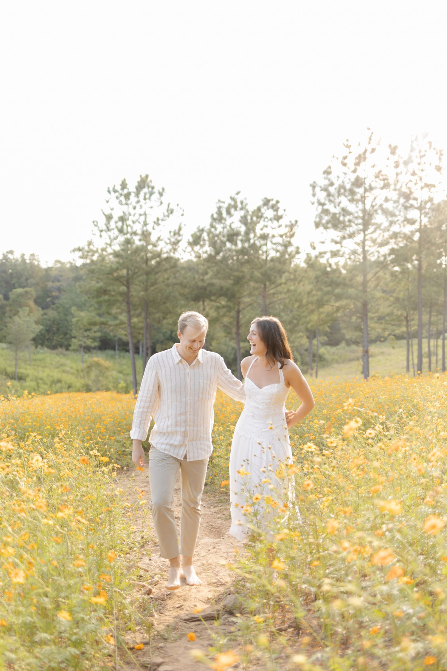 Couple in white outfits walking hand in hand through a blooming yellow wildflower field for engagement photoshoot in Chapel Hill, NC.