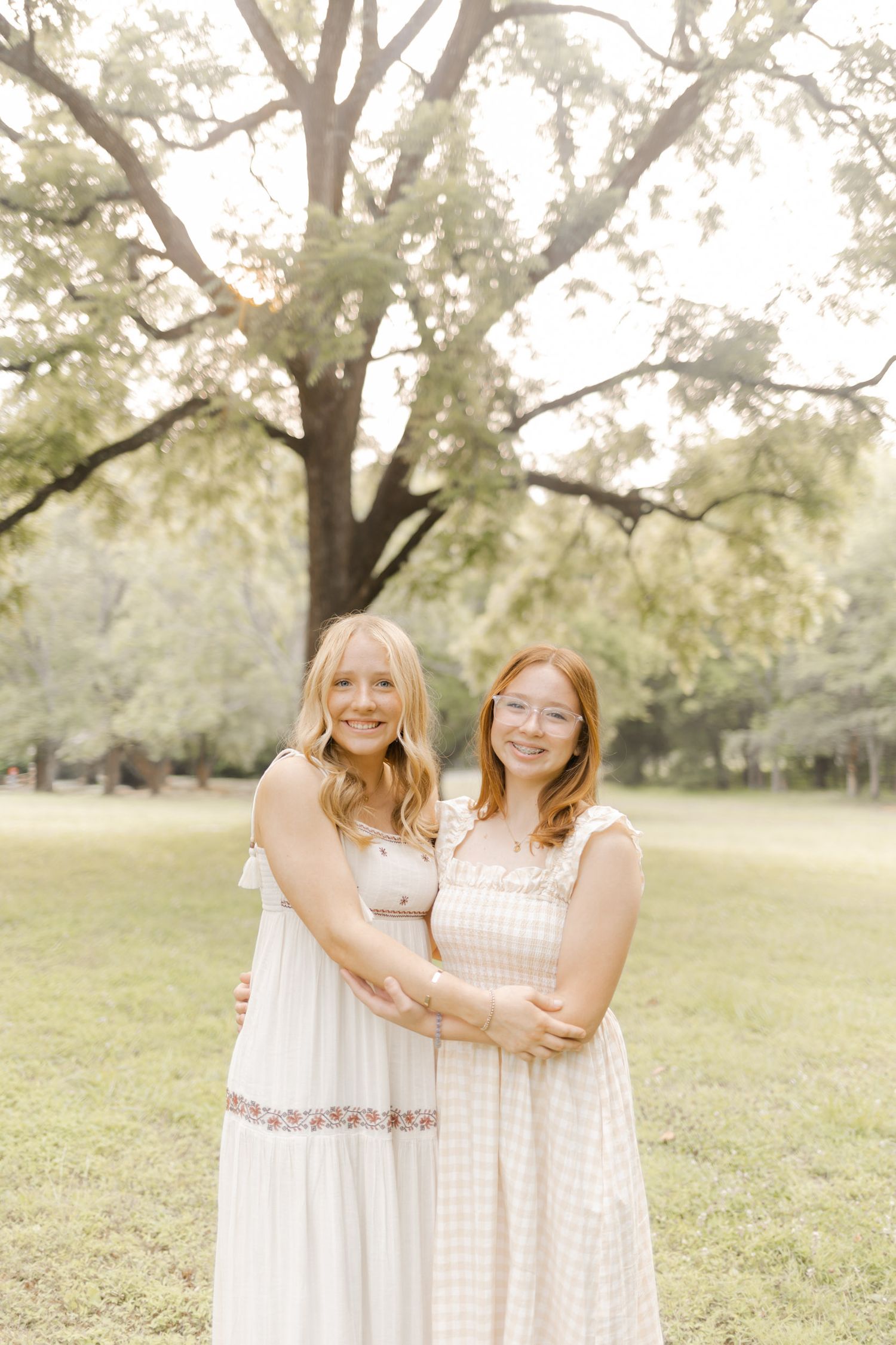 Friends in coordinating white dresses share joyful moments under a large tree.