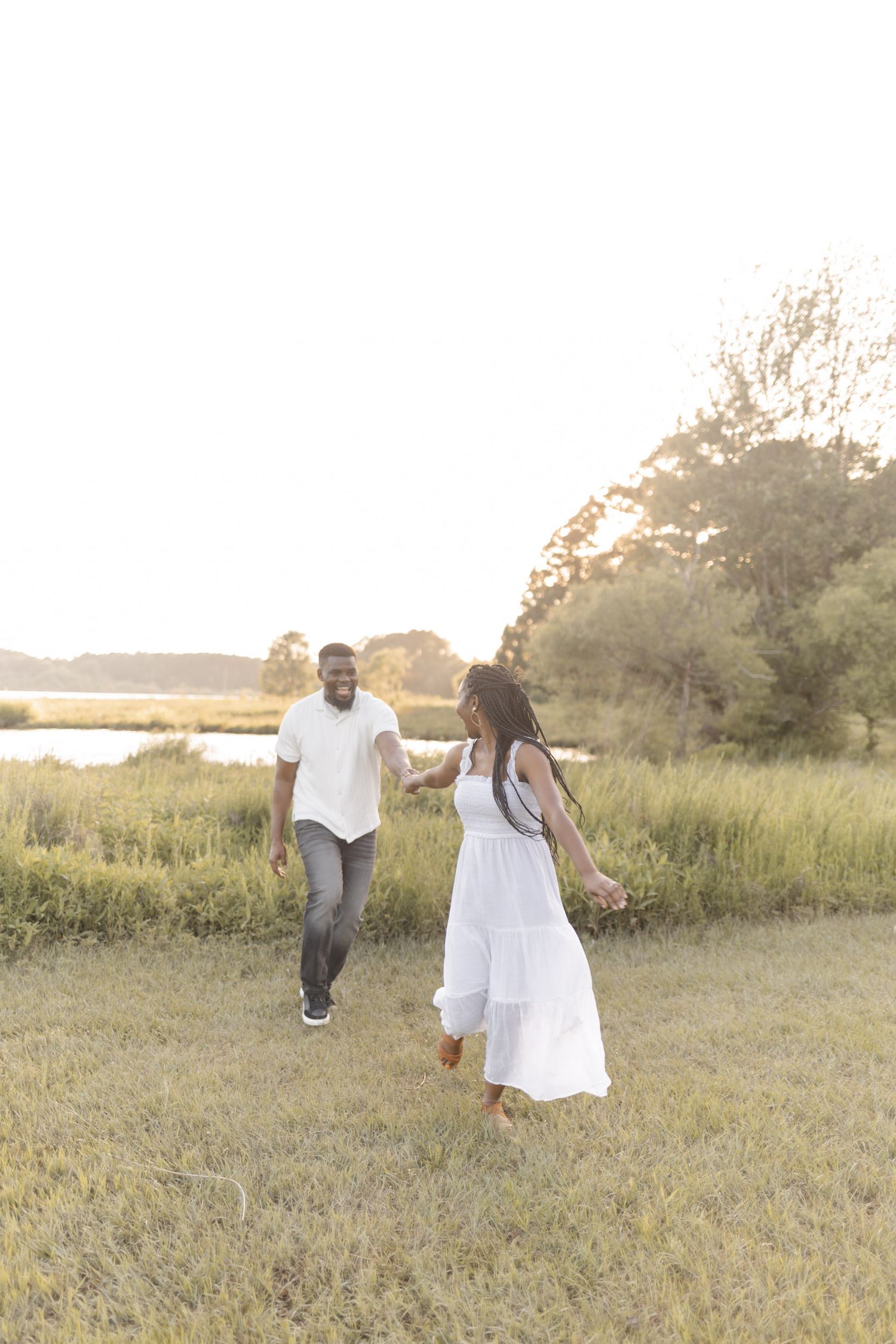 A couple walks hand-in-hand through a sunlit field, the woman in a white dress and the man in a white shirt and jeans.