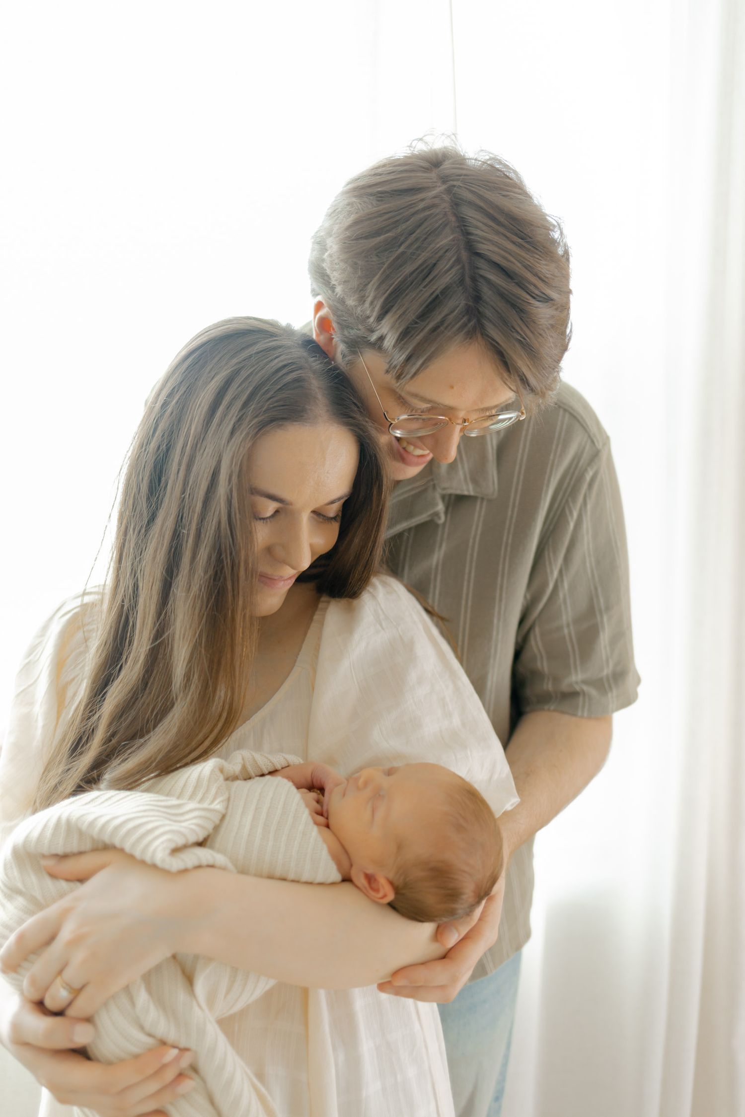 Parents lovingly gaze at their newborn baby wrapped in white, sharing a tender family moment in soft natural light.