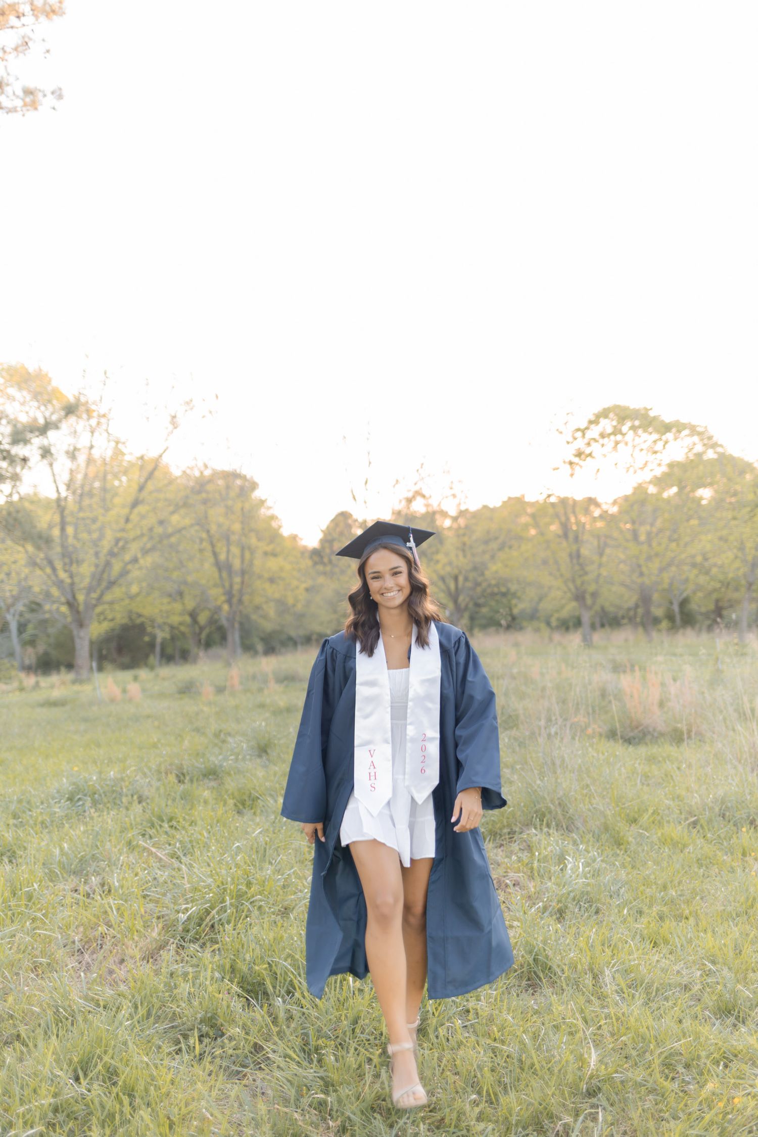 Smiling graduate in navy cap and gown walking through green field at sunset.