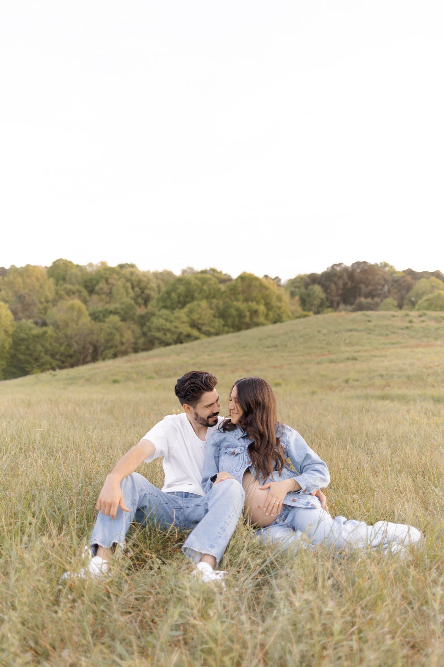 Couple sitting in a grassy meadow, sharing a romantic moment, dressed in casual denim outfits with trees in the background.