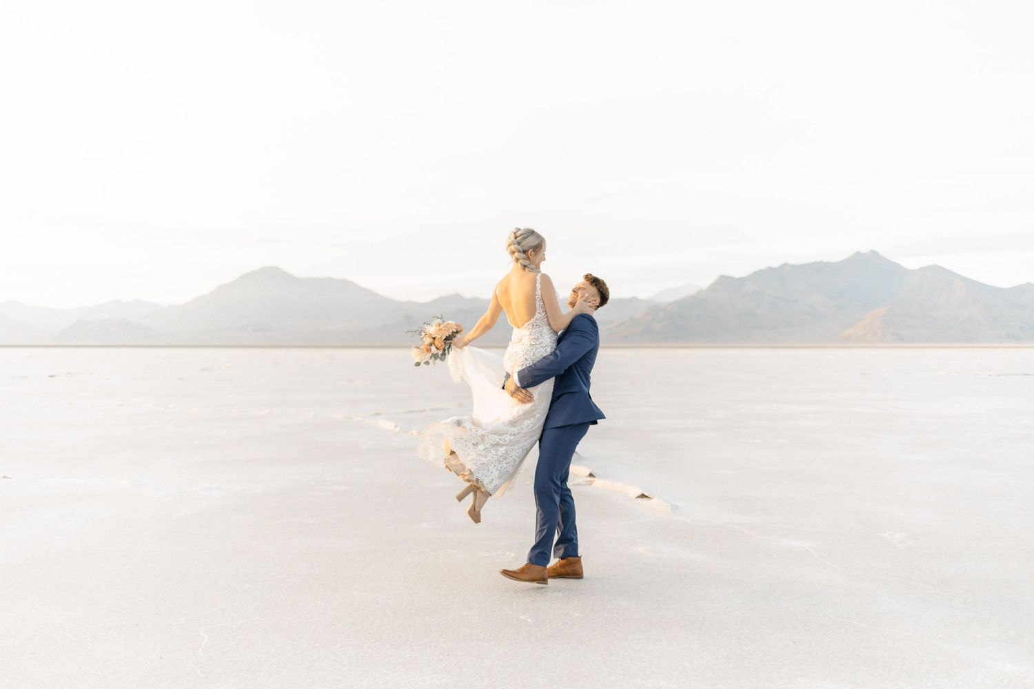 Couple in formal wedding attire dancing together on a bright white beach landscape with mountains in background.