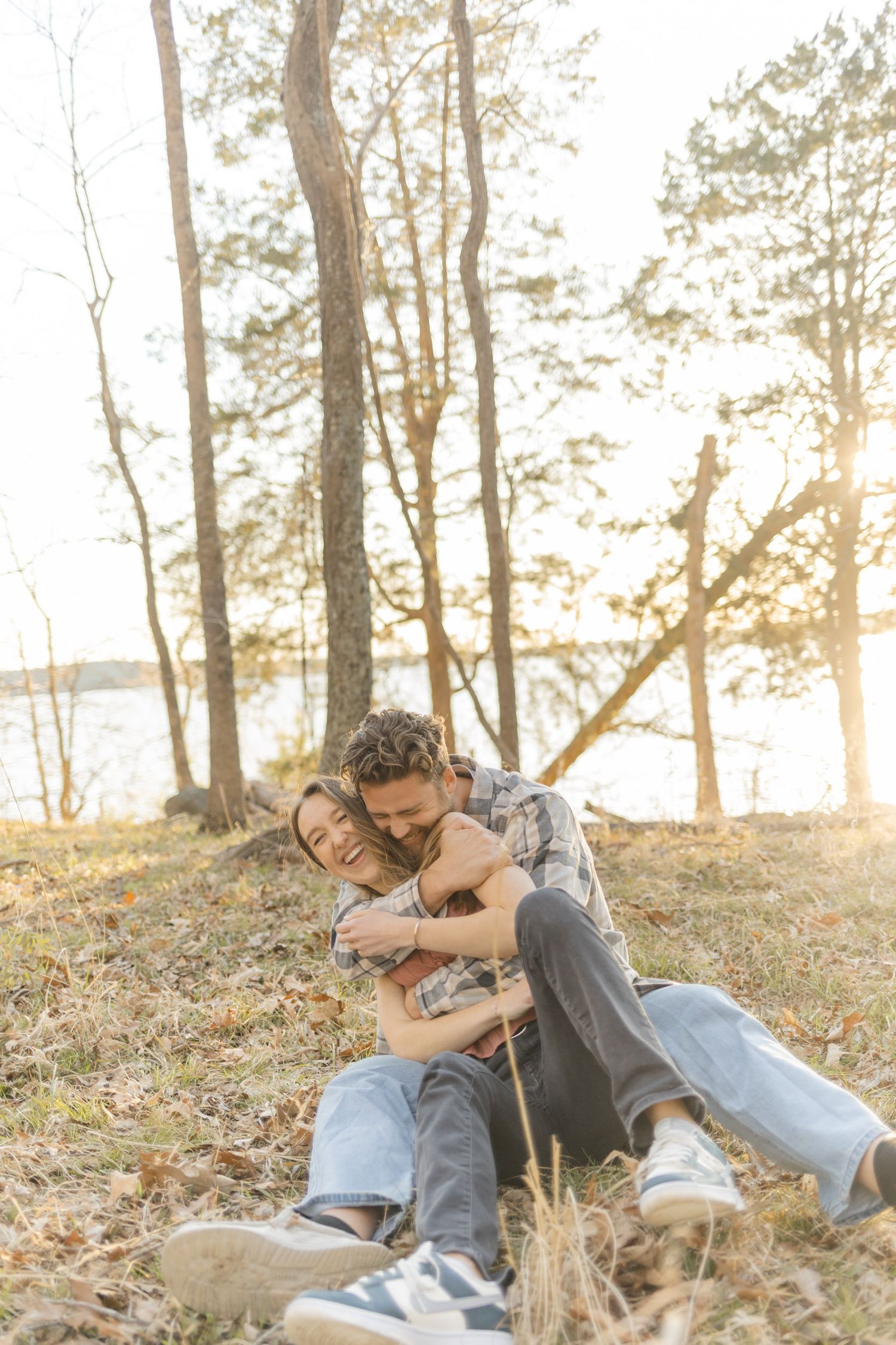 A couple sharing a playful moment on fallen leaves with sunlit trees in the background during their couples lifestyle photoshoot in Raleigh,