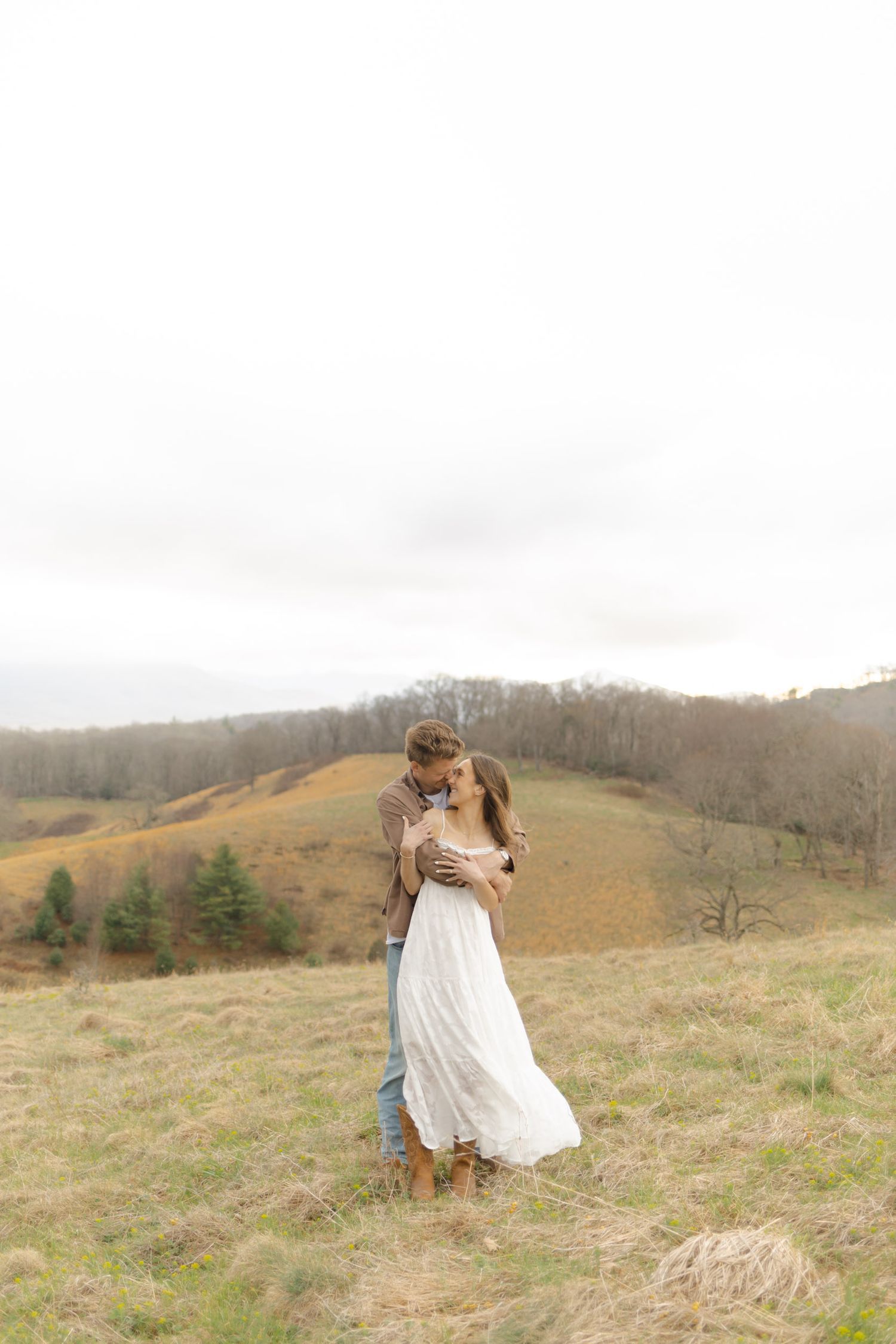 Couple embracing on a scenic hillside meadow, woman in flowing white dress and man in brown jacket, rolling hills backdrop.