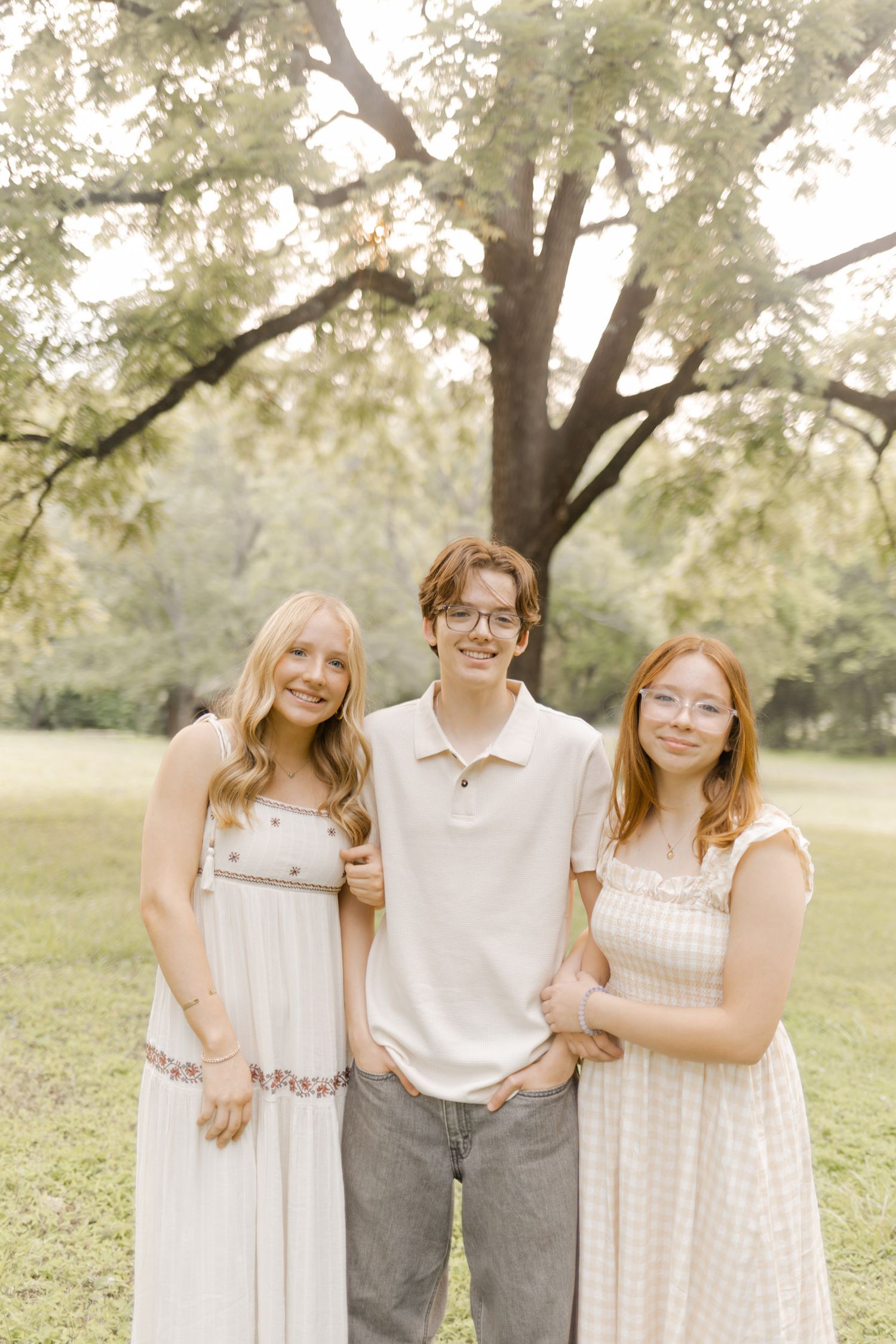 Three people pose together outdoors wearing coordinated light colored casual outfits while doing a photoshoot.