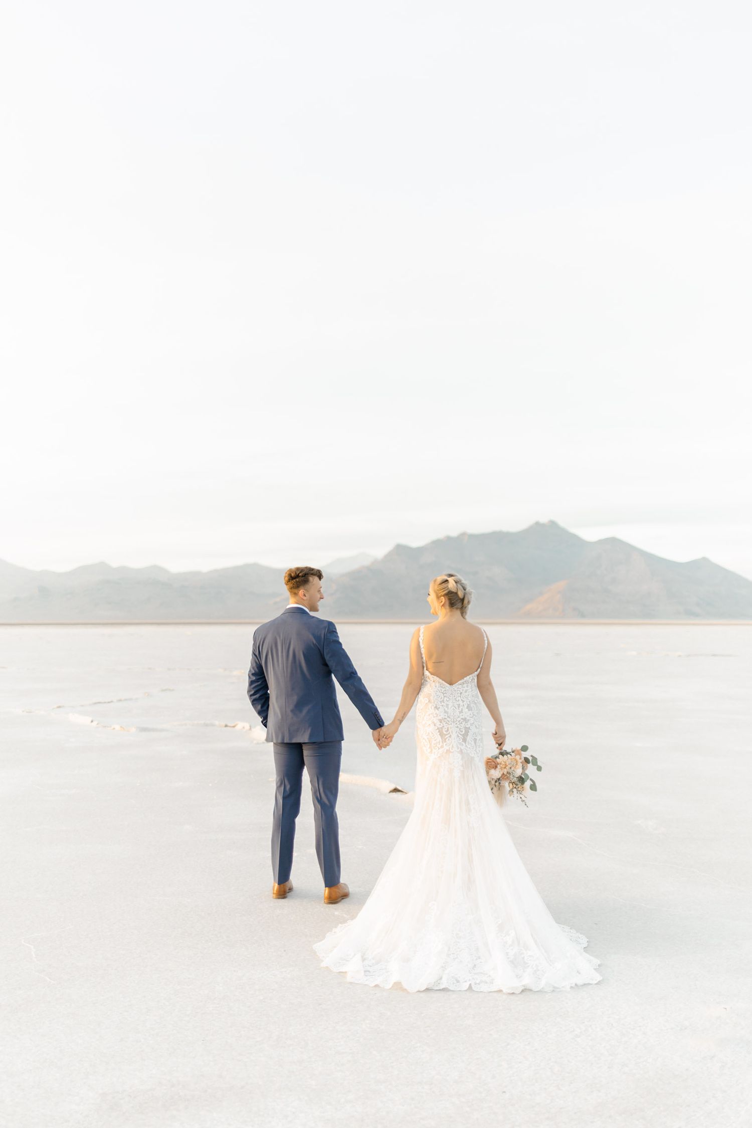 Bride and groom holding hands on the Bonneville Salt Flats, bride in lace gown with bouquet, mountains in background.