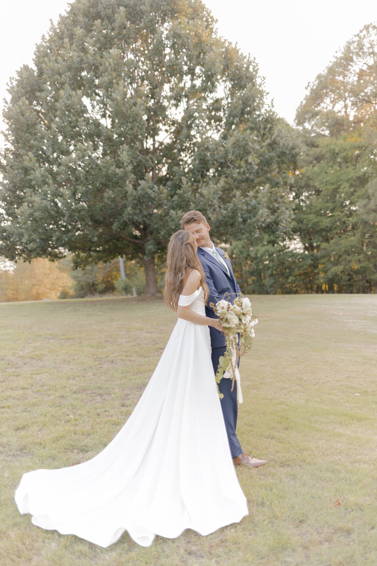 Bride in white gown with train and groom in navy suit embrace outdoors near large trees on their wedding day.
