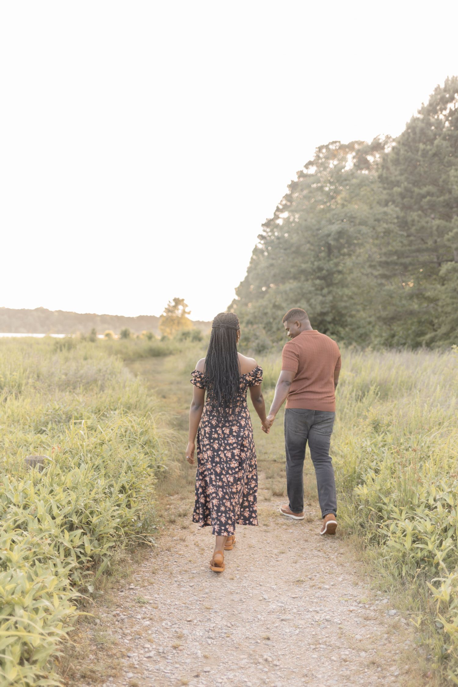 Couple holding hands walking down a scenic path surrounded by lush green fields and trees at golden hour.