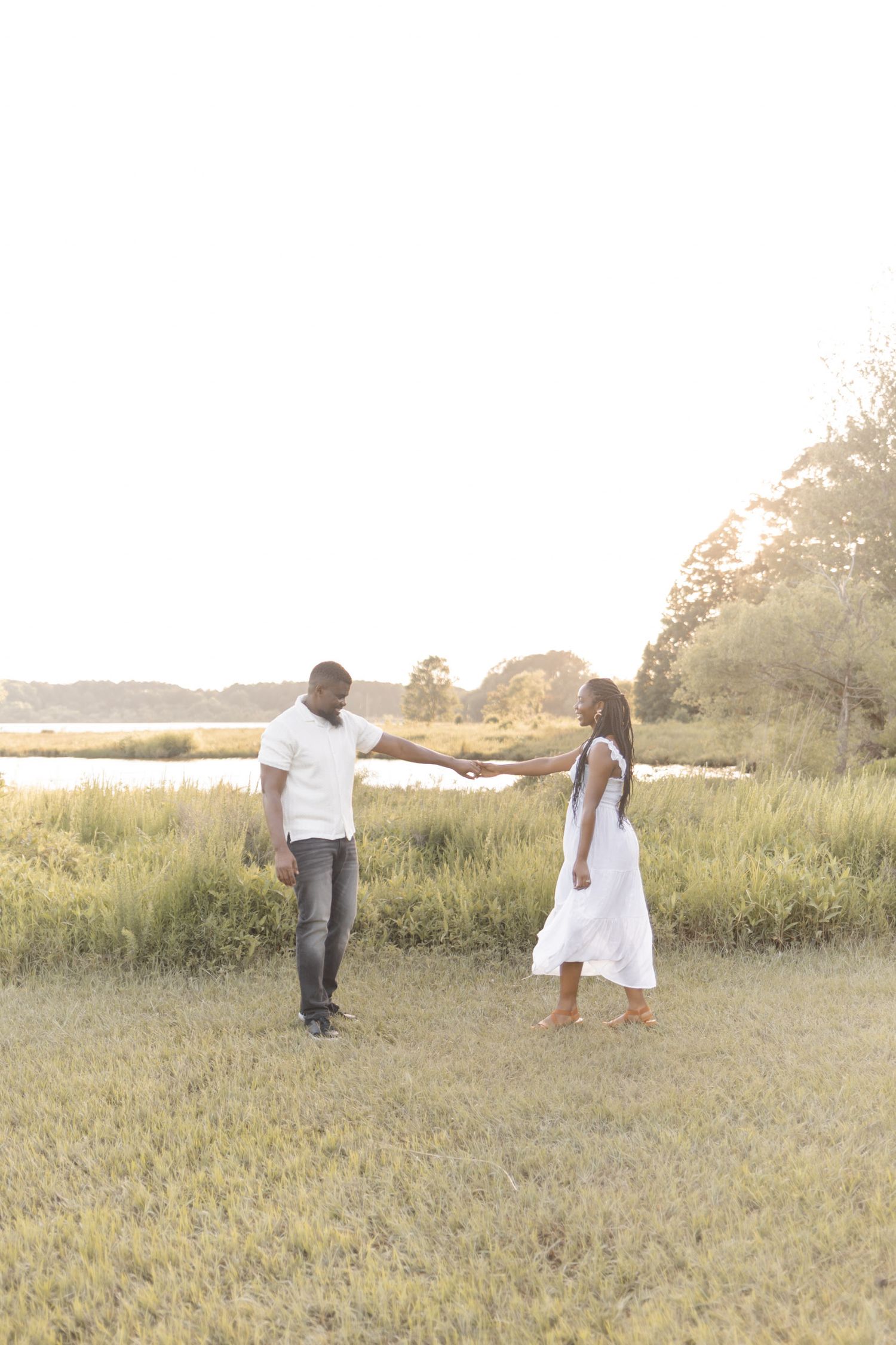 A couple in white outfits holds hands in a sunlit grassy field near a serene waterway surrounded by trees.