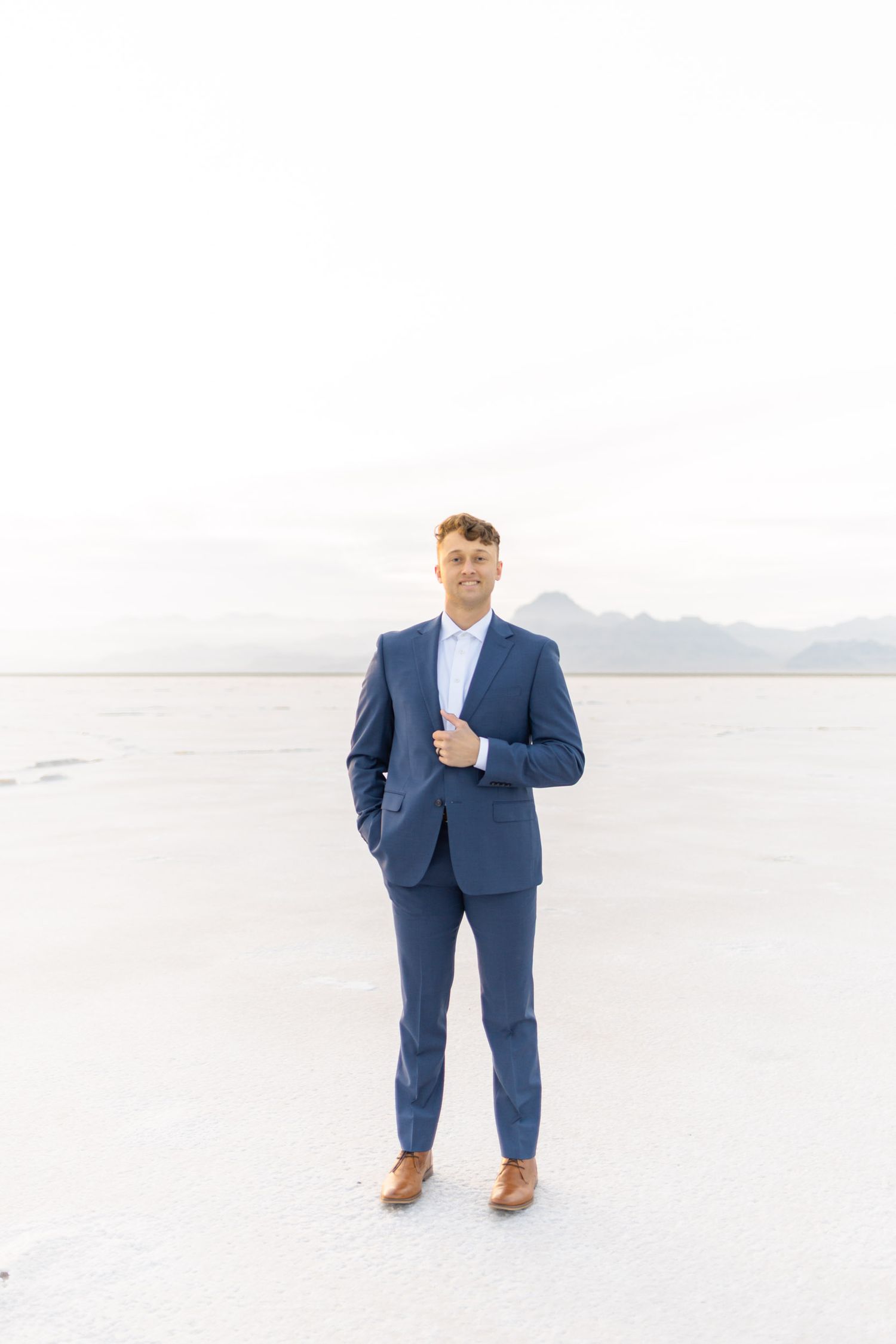 A groom in a navy blue suit stands on a white salt flat landscape with mountains in the background.