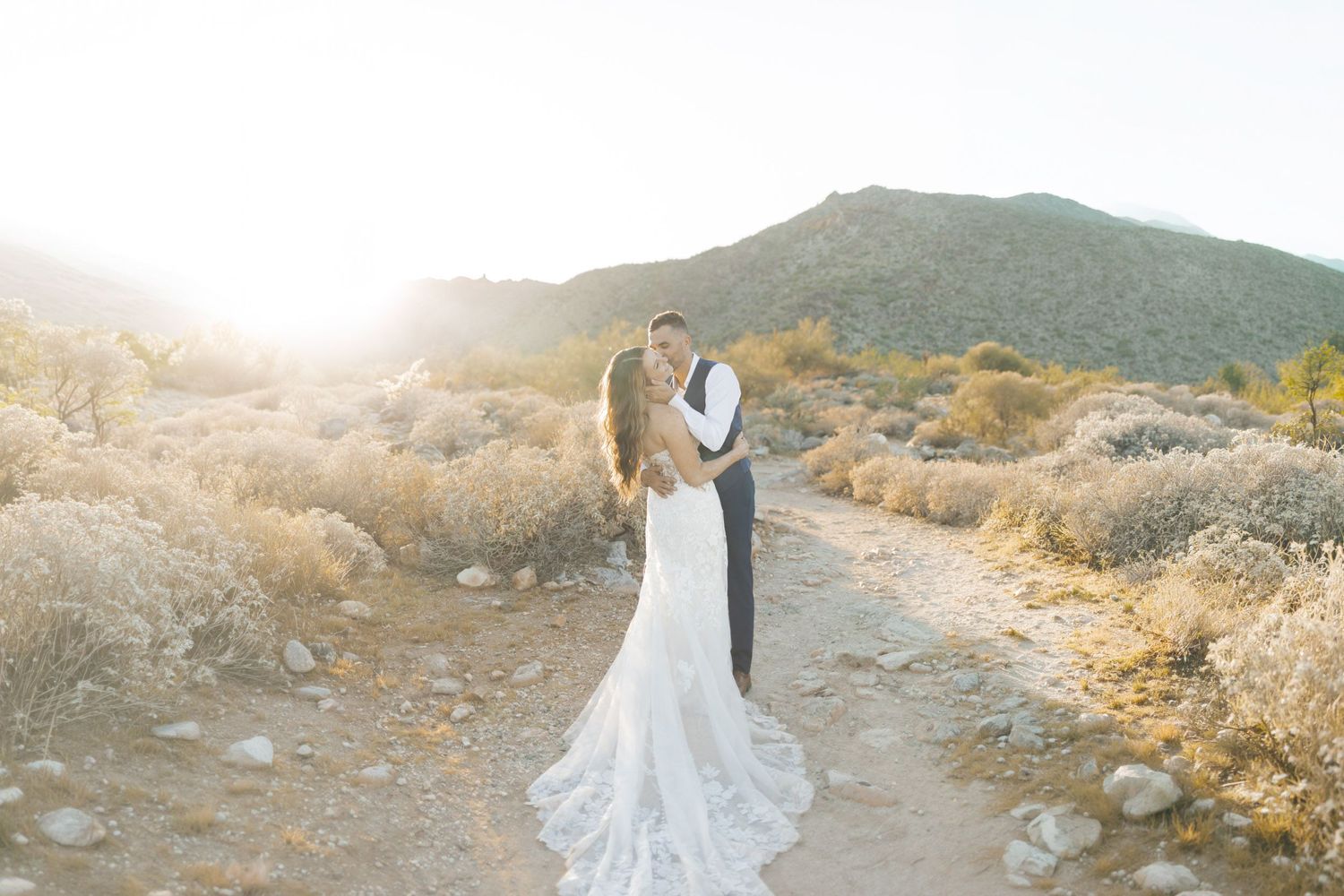 Bride and groom embrace on a desert path with golden sunlight and mountain backdrop on their wedding day.