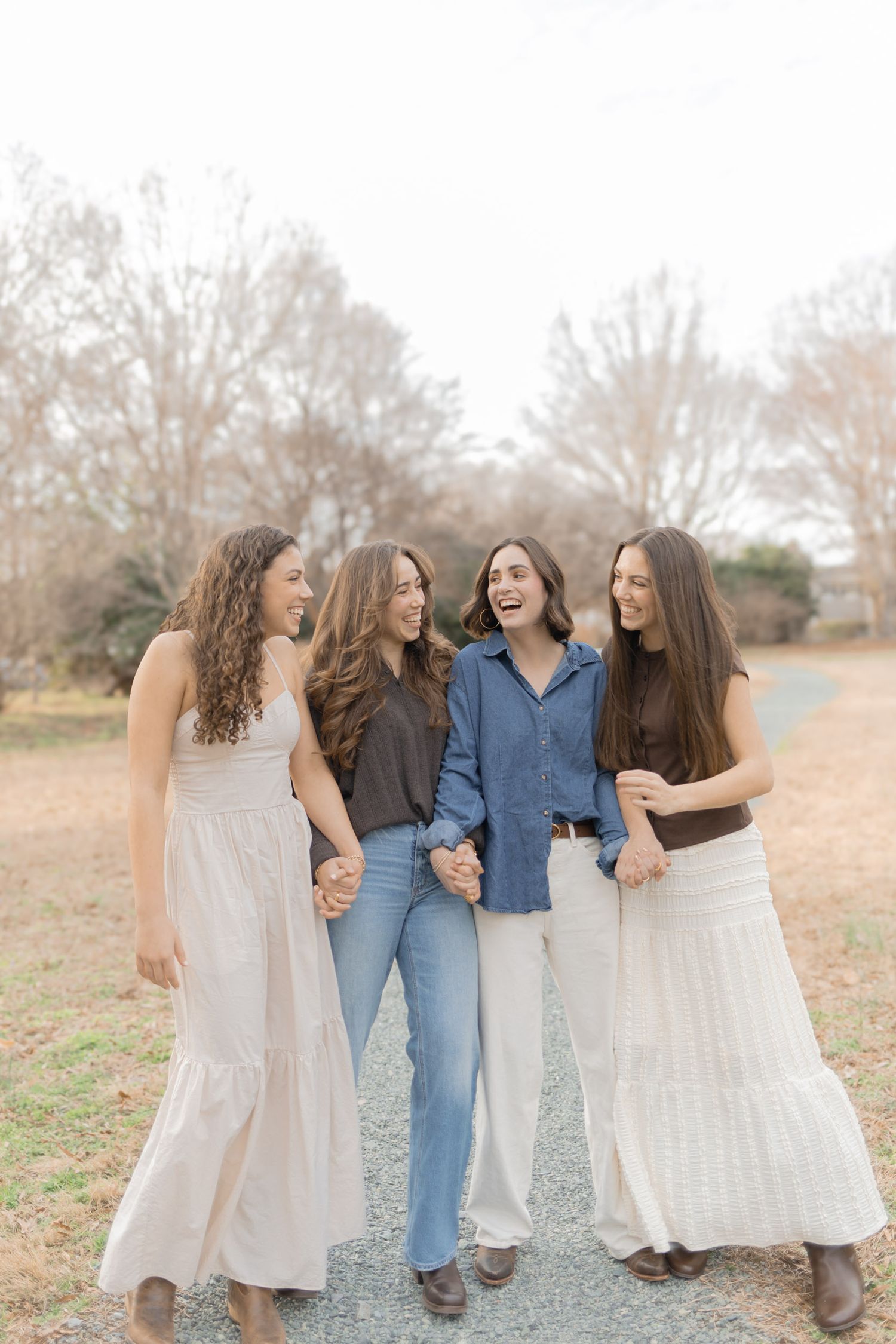 Four laughing young women walking together on a gravel path in casual outfits.