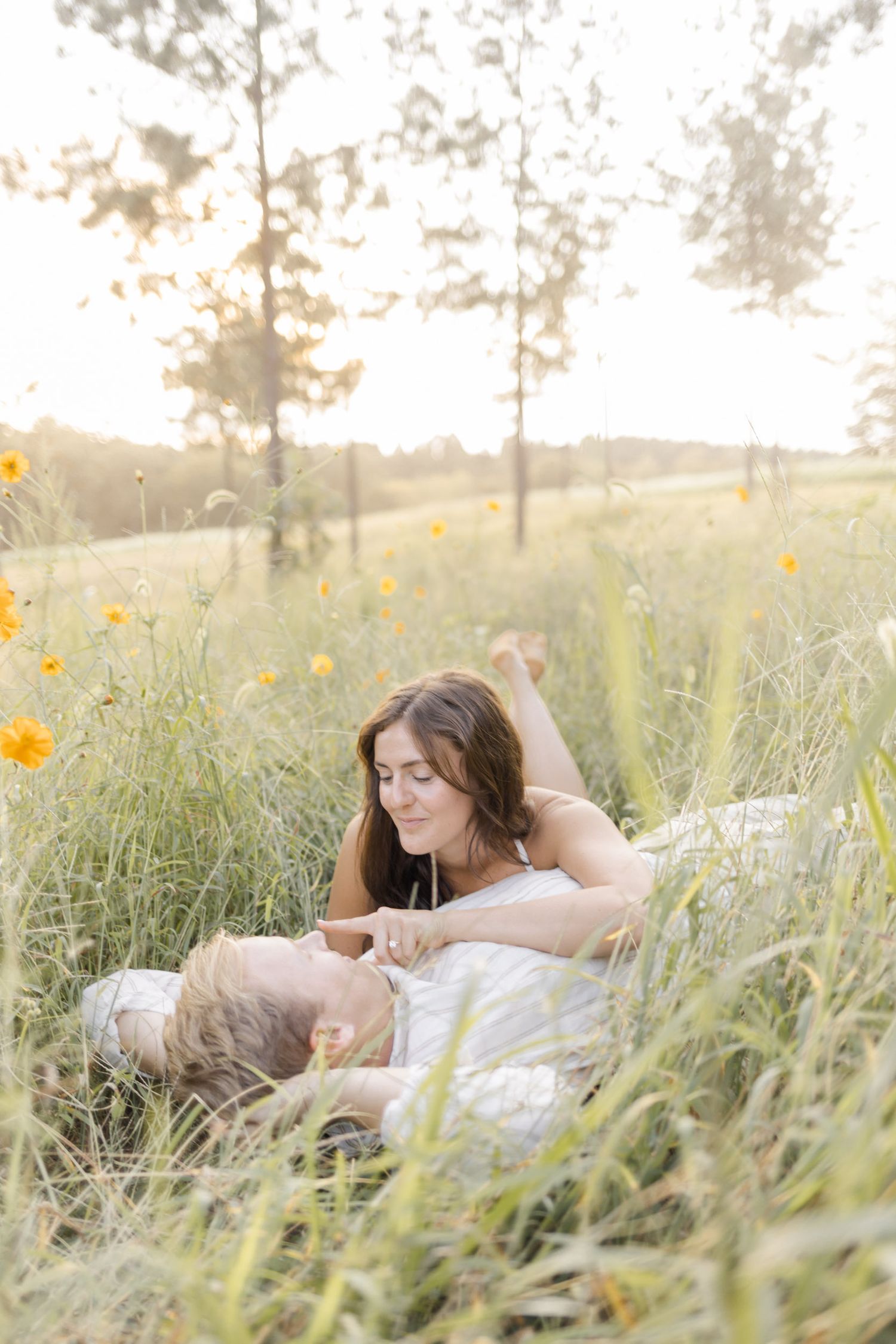 Two people lying together in tall grass with yellow flowers at sunset during engagement session with photographer in North Carolina.