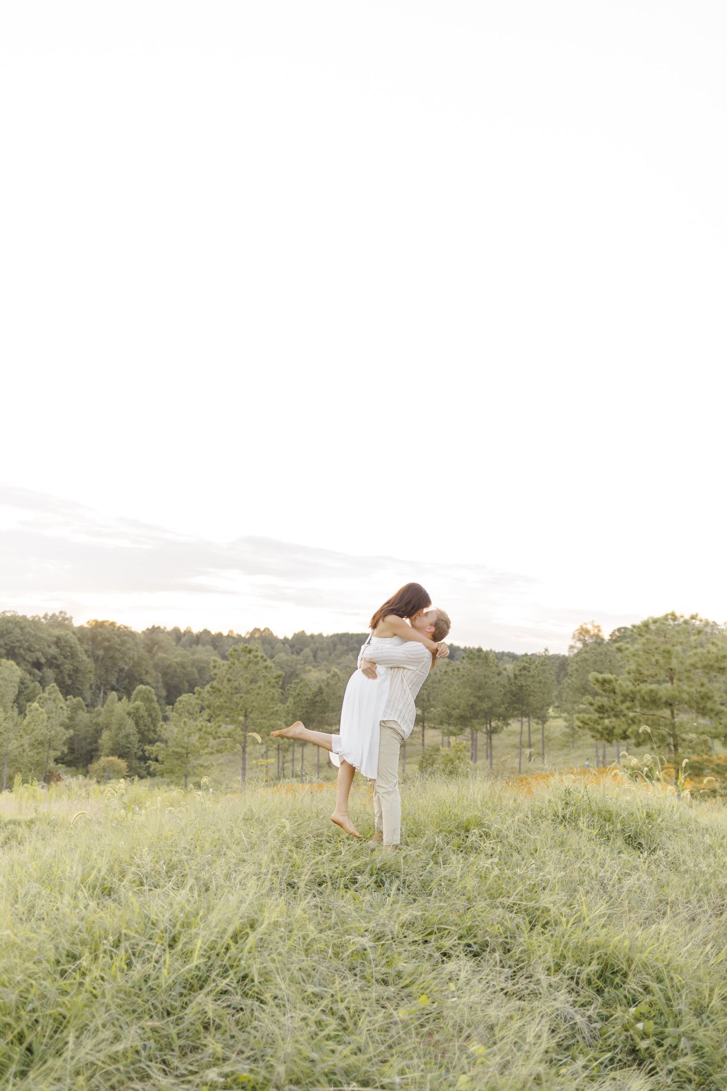 A romantic couple embraces in a sunlit meadow with rolling hills and trees in the background during engagement session in Chapel Hill, NC.