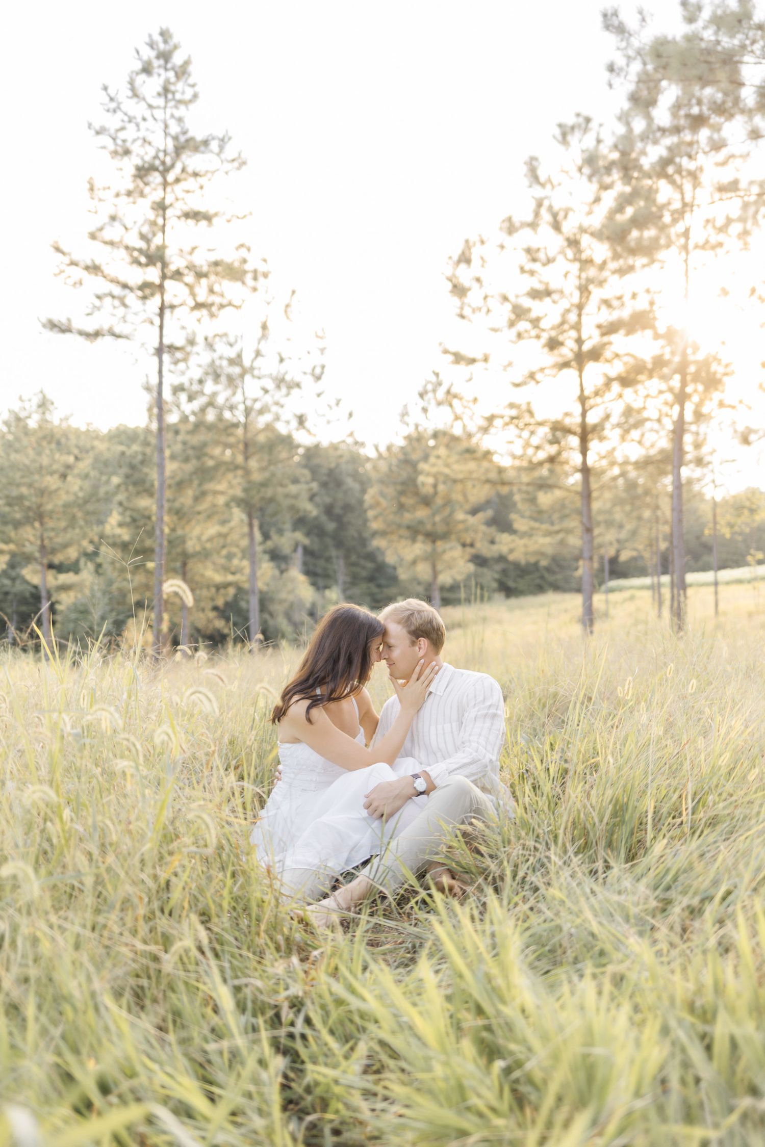 A couple in white clothing sits together intimately in a sunlit meadow with tall pine trees in the background at photoshoot in Chapel Hill