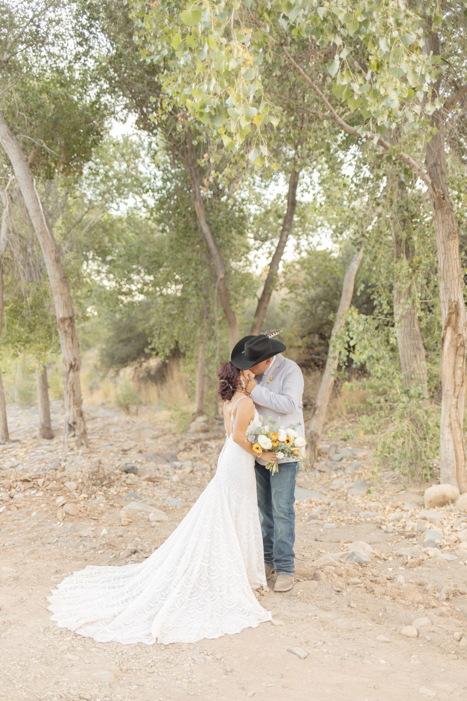 Bride and groom kissing in a rustic outdoor setting, bride in lace gown holding yellow bouquet, groom in cowboy hat.