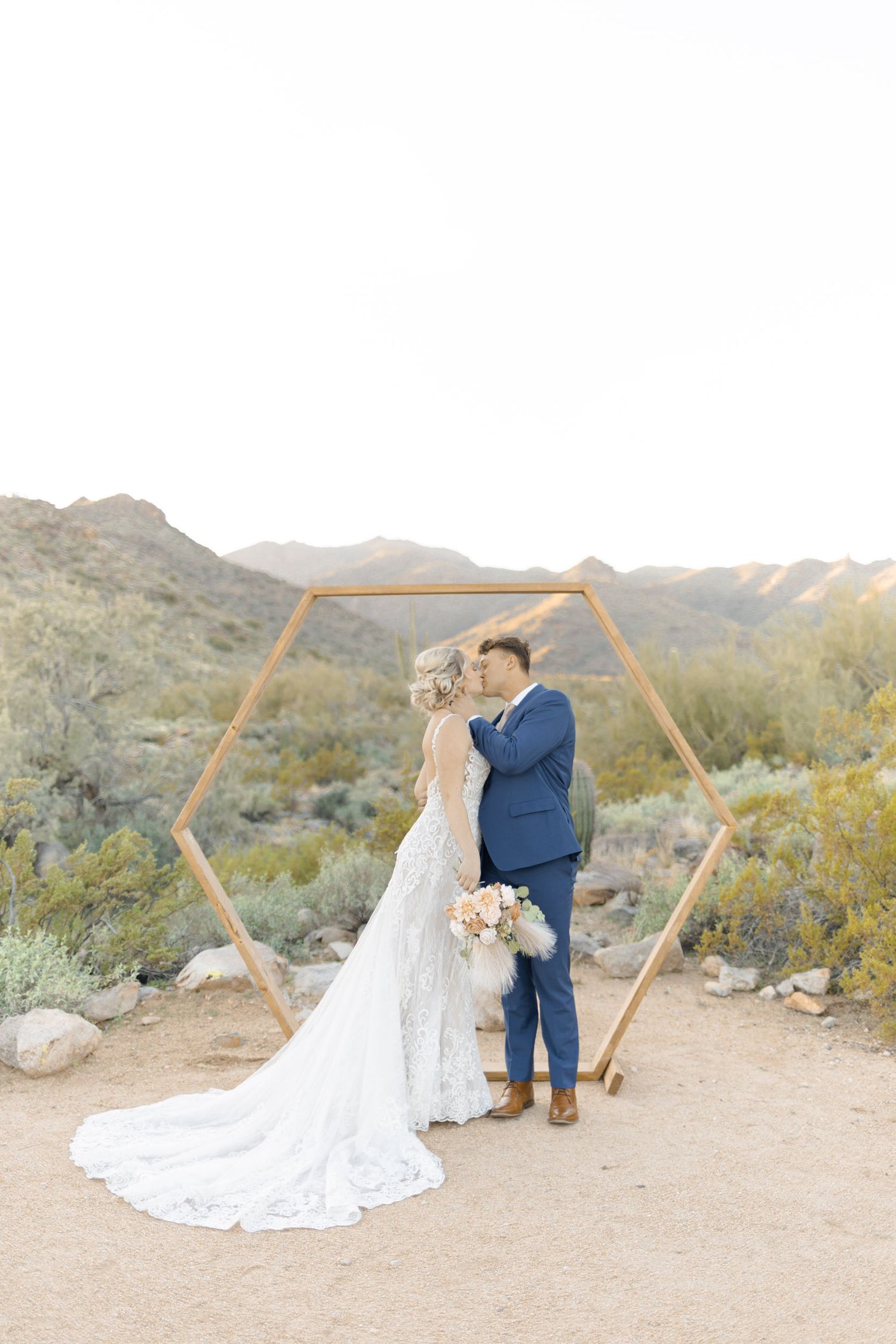 Bride and groom kissing under a hexagonal wooden arch in the Arizona desert with mountain views.