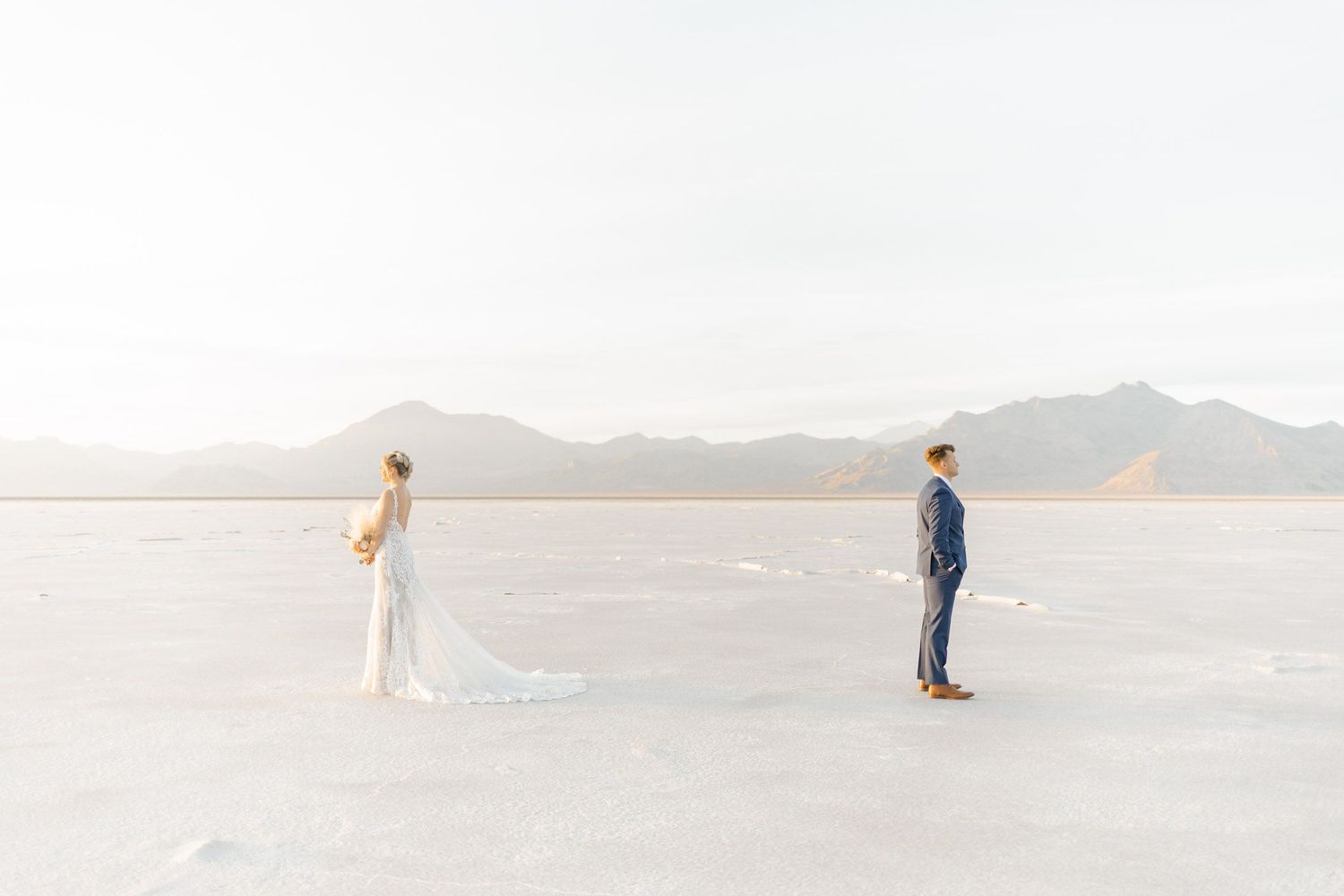 Bride and groom stand apart on vast white salt flats with mountain backdrop during desert elopement ceremony.