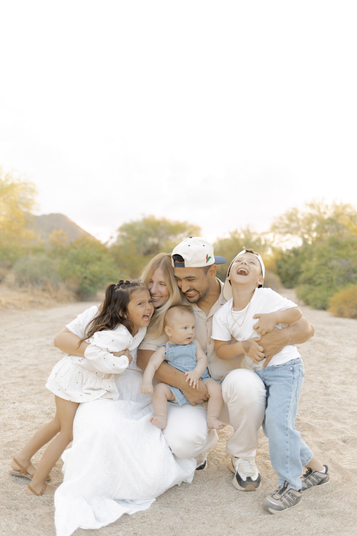 A happy family of five with three children laugh and cuddle together on a sandy desert path.