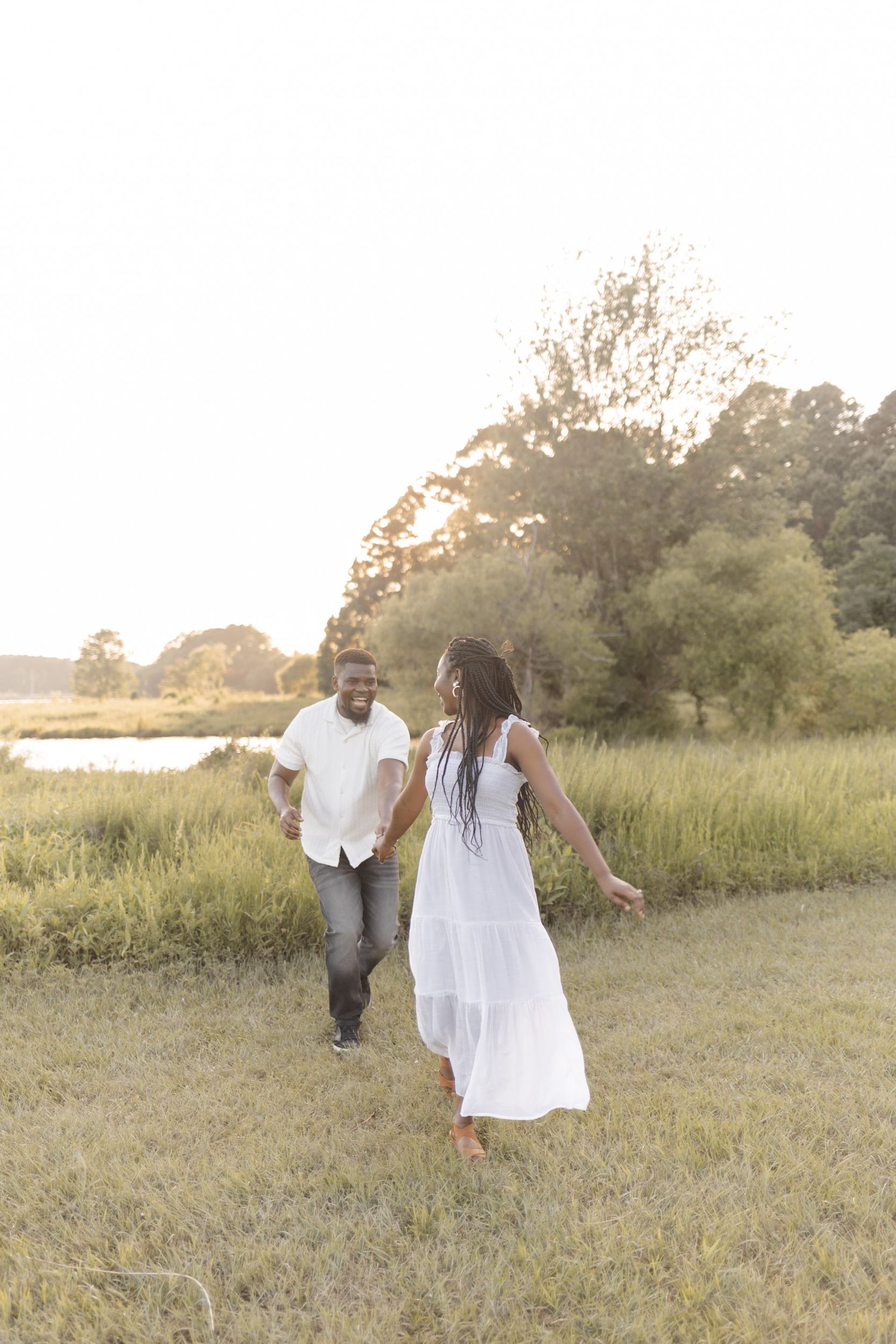 A couple walks hand-in-hand through a sunny green meadow, the woman wearing a flowing white dress.