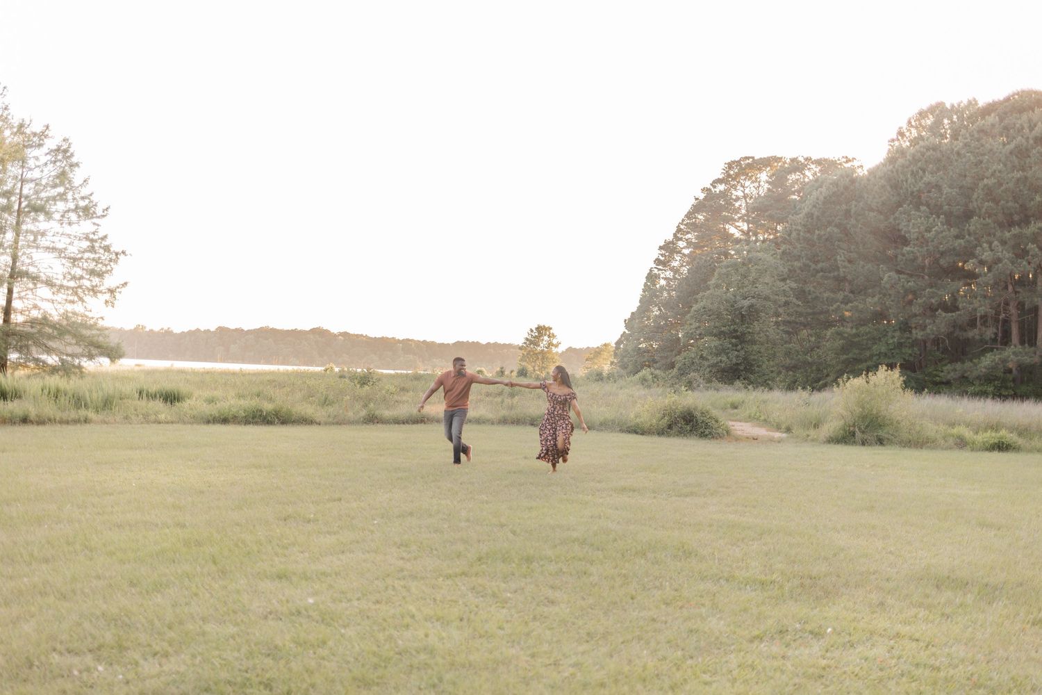 A joyful Black couple runs hand-in-hand across a lush green field with trees and a lake in the golden background.