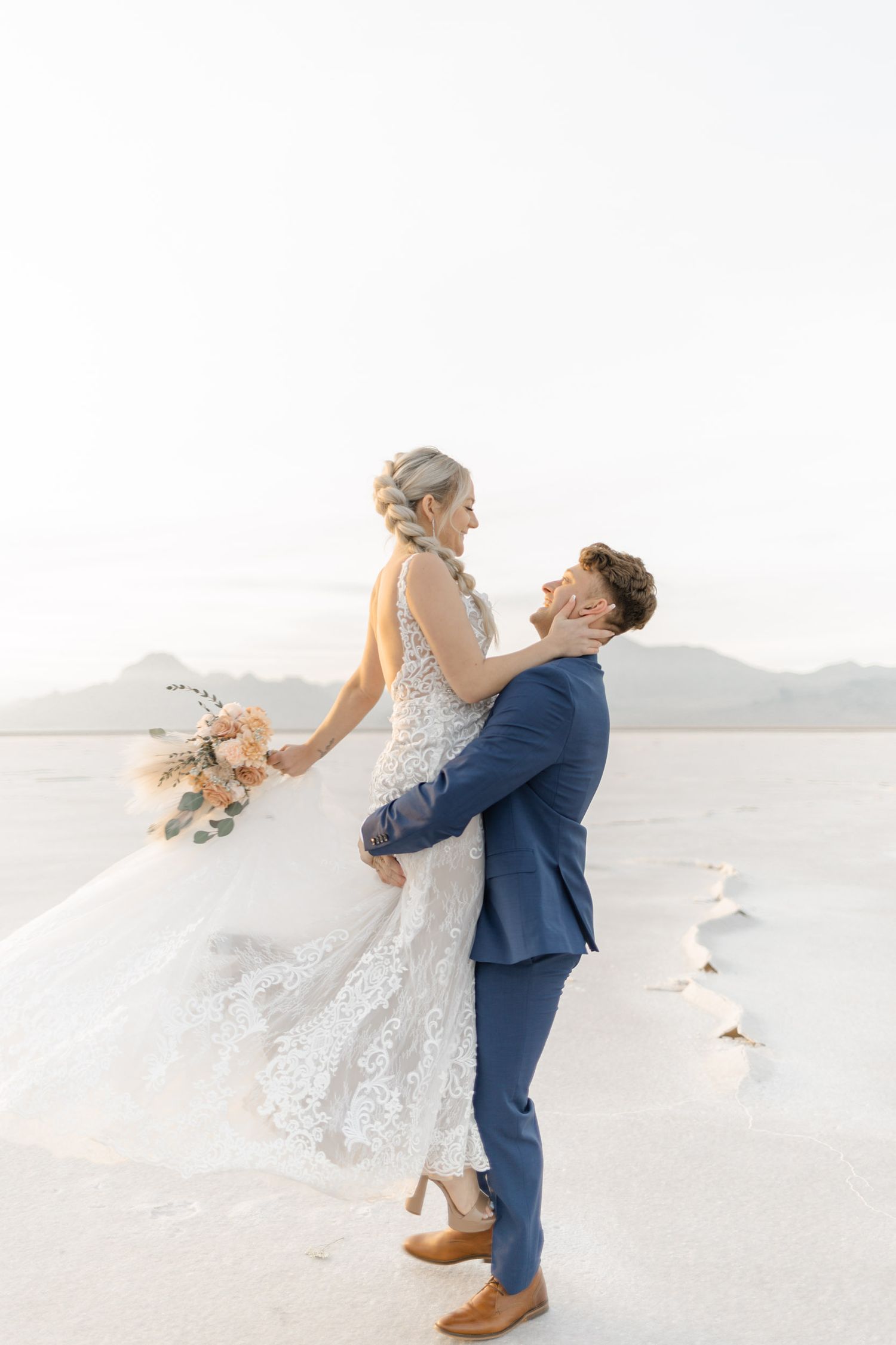 A romantic wedding couple in white dress and navy suit share a joyful moment on a bright salt flat landscape.