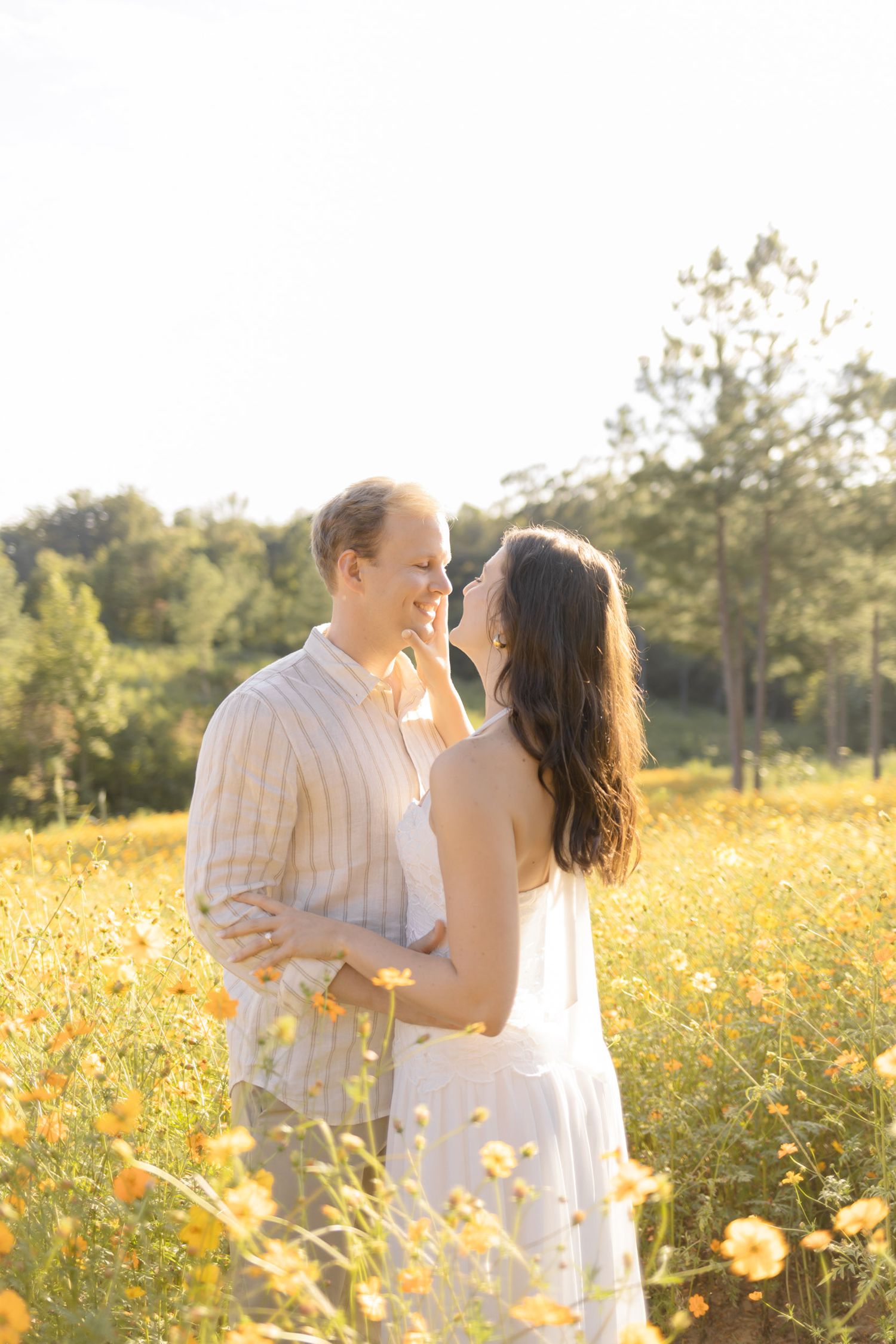 Tender moment between a couple in white clothing amid yellow flowers and evening sunlight for engagement photoshoot in Chapel Hill, NC.
