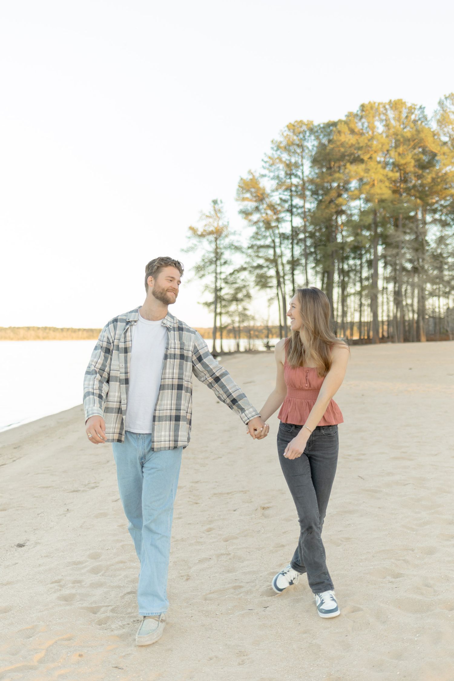Playful couple walking and laughing together on sandy slope with pine forest backdrop in Raleigh, North Carolina.