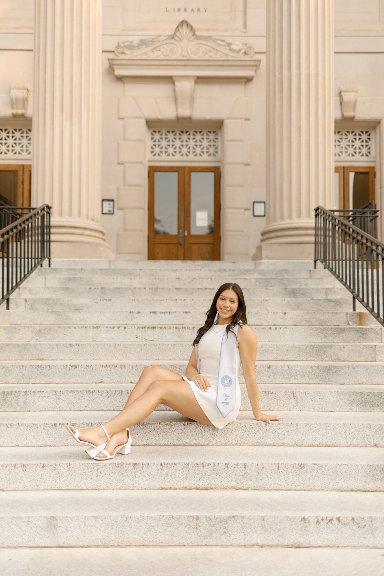 Graduate in white dress with honor stole sitting on library steps for senior photos.