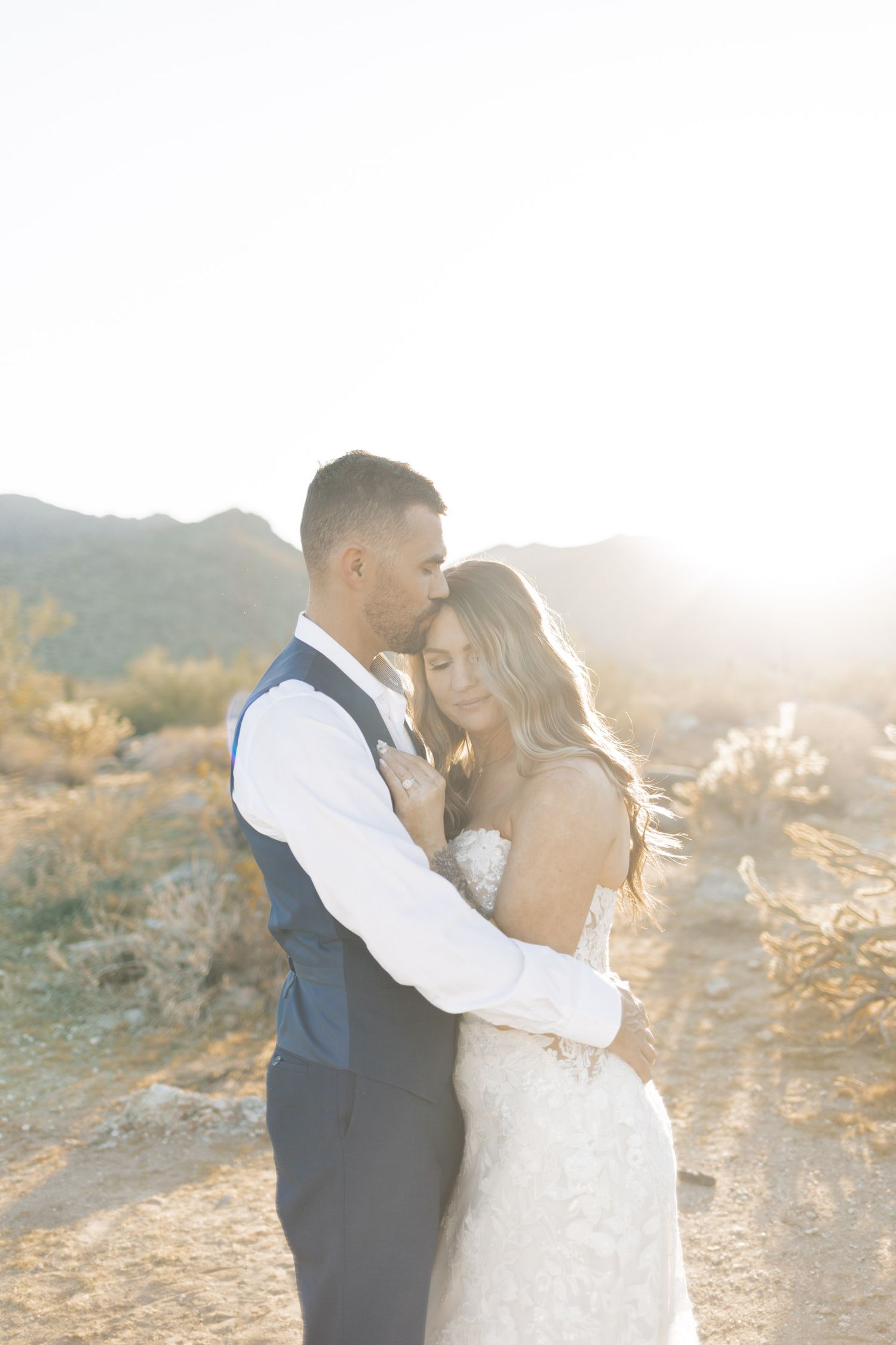 Couple embracing in golden desert sunset light, bride in lace gown and groom in navy suit, mountains in background.