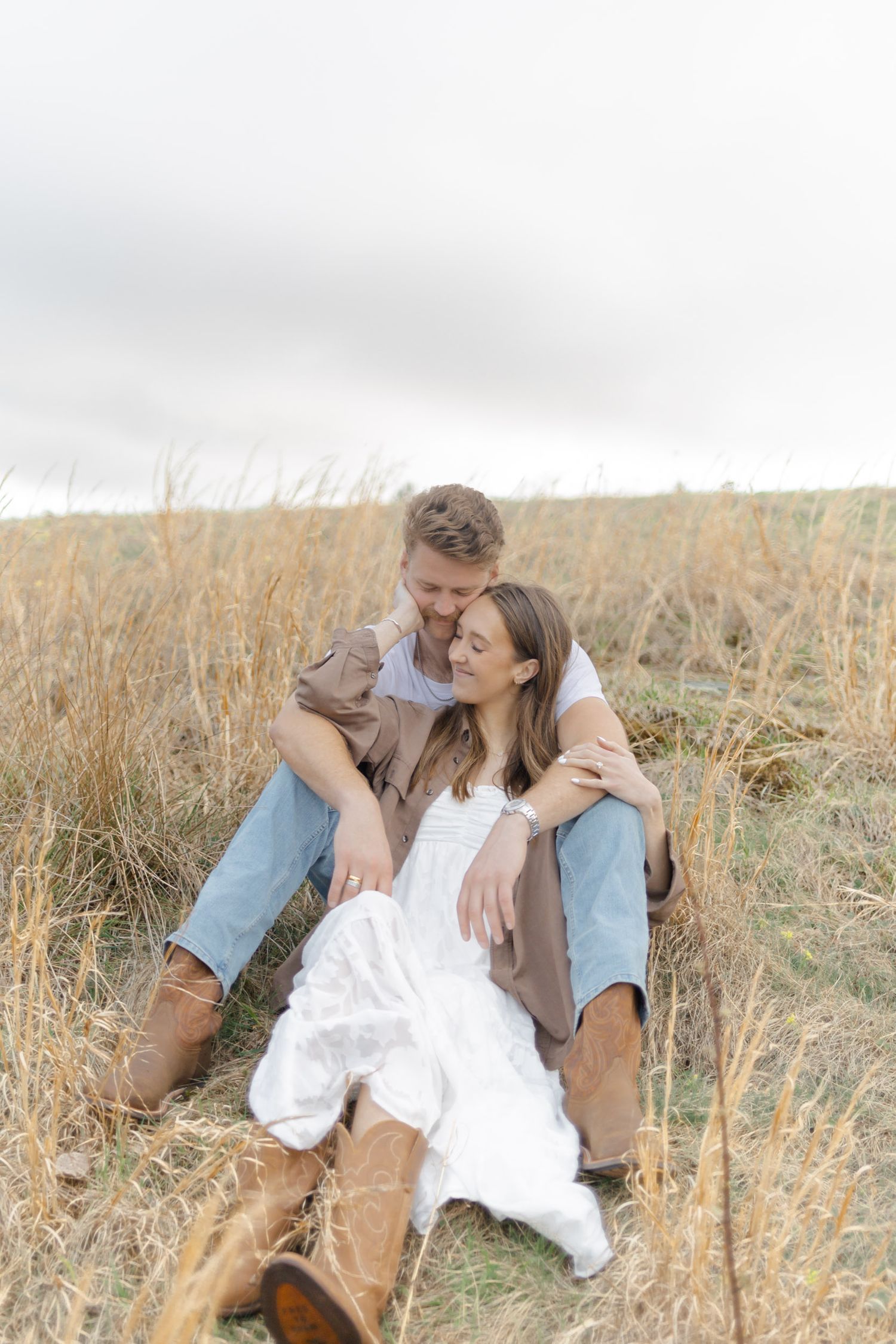 Couple sitting in golden grass field, man embracing woman in white dress and cowboy boots.