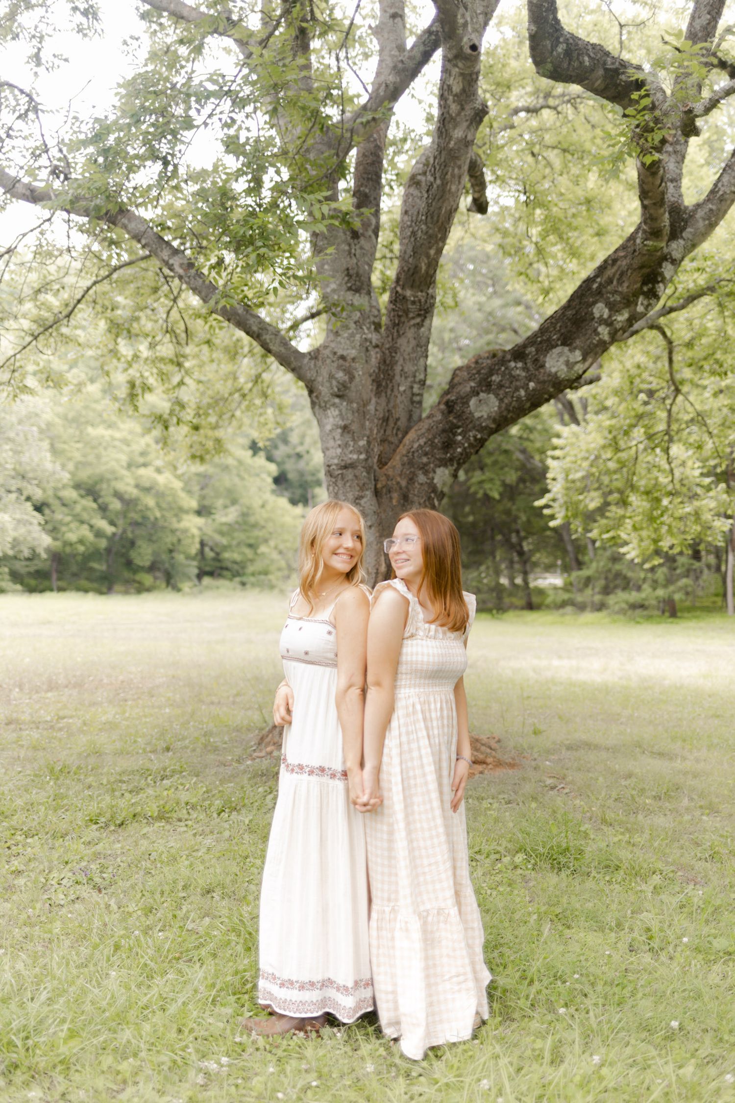 Two people in flowing cream dresses stand together in a sunlit meadow beneath large trees in Durham, North Carolina.