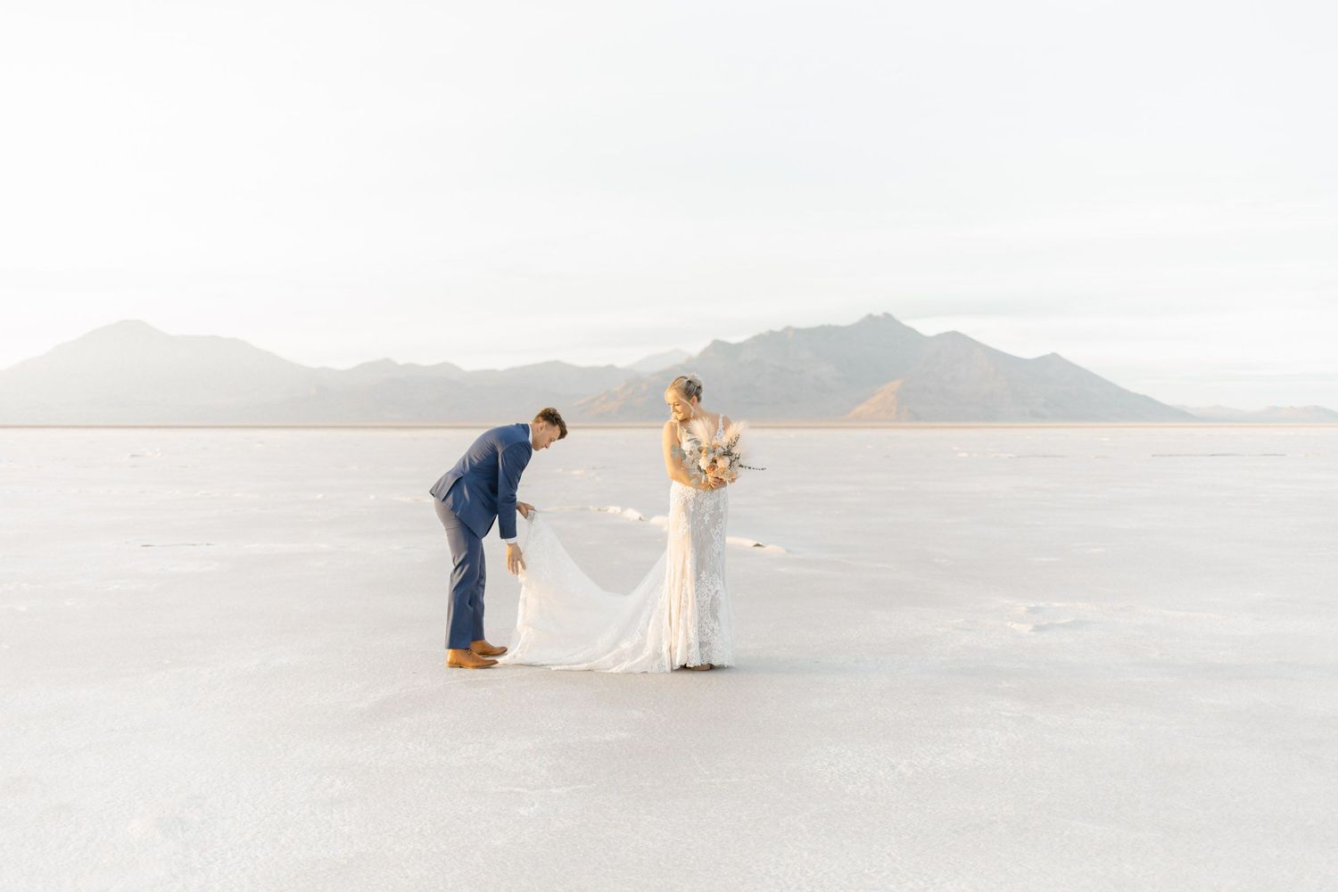 Couple in elegant wedding attire poses on vast salt flats with mountains in background during sunset photoshoot.