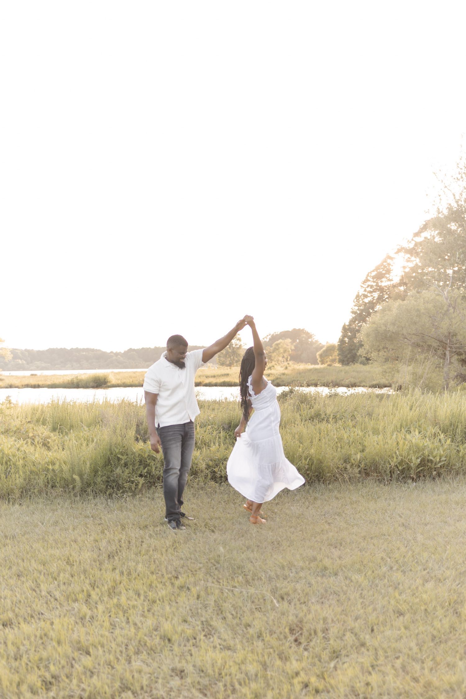 Man spinning woman in white dress while dancing in a golden sunlit field near a serene lake at sunset.