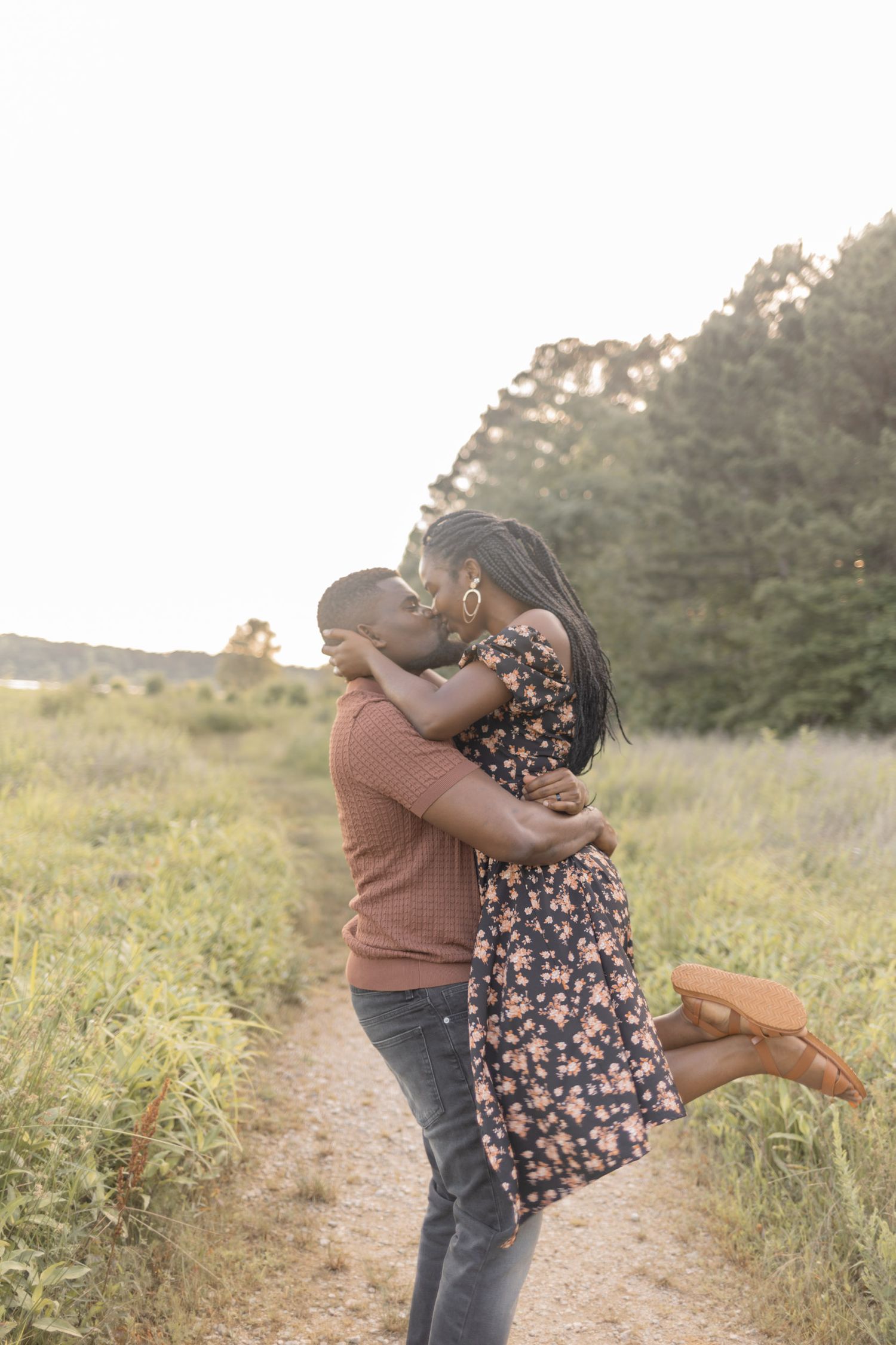Couple sharing a romantic kiss on a scenic field path at golden hour, woman lifted off ground in floral dress.